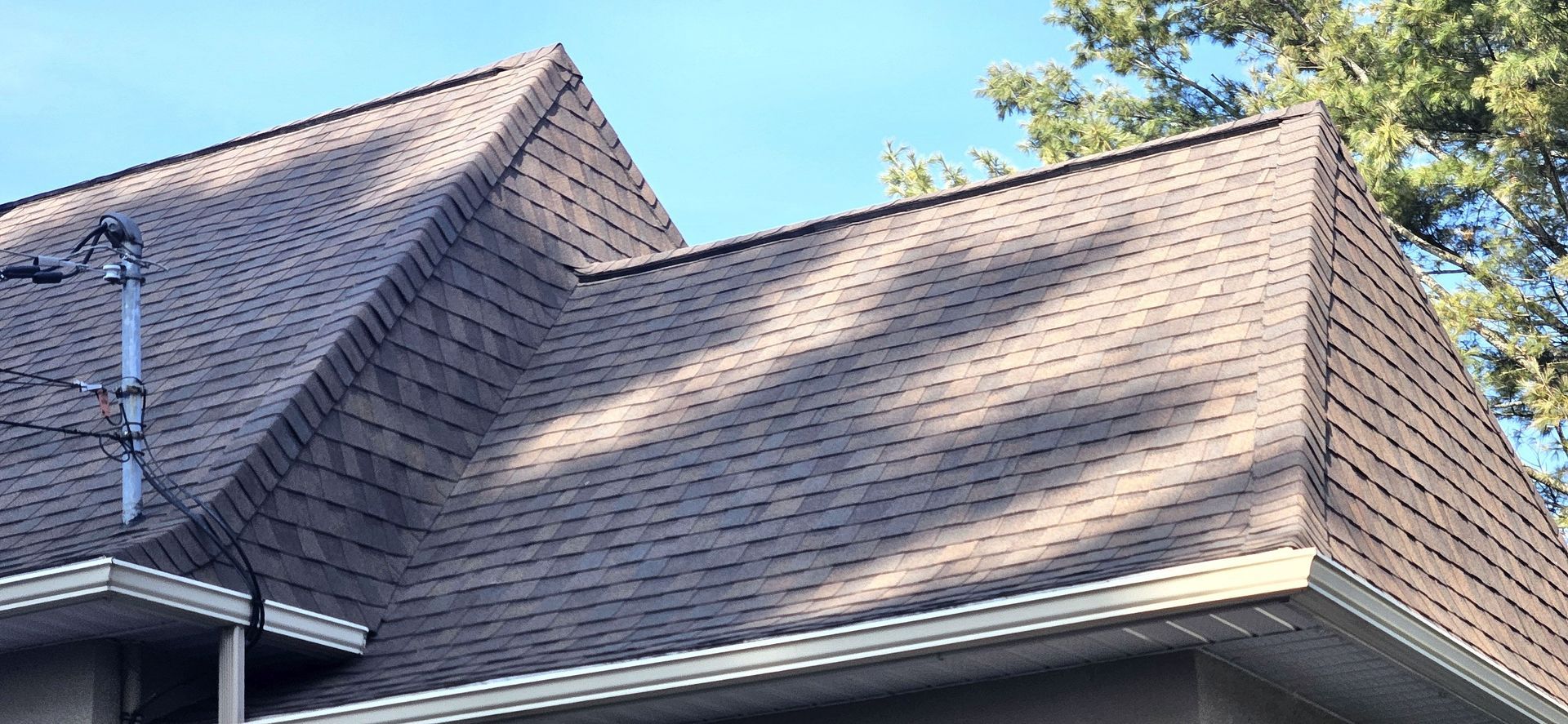 Gros plan sur un toit de bardeaux bruns sous un ciel bleu clair, avec les ombres projetées par les arbres environnants.