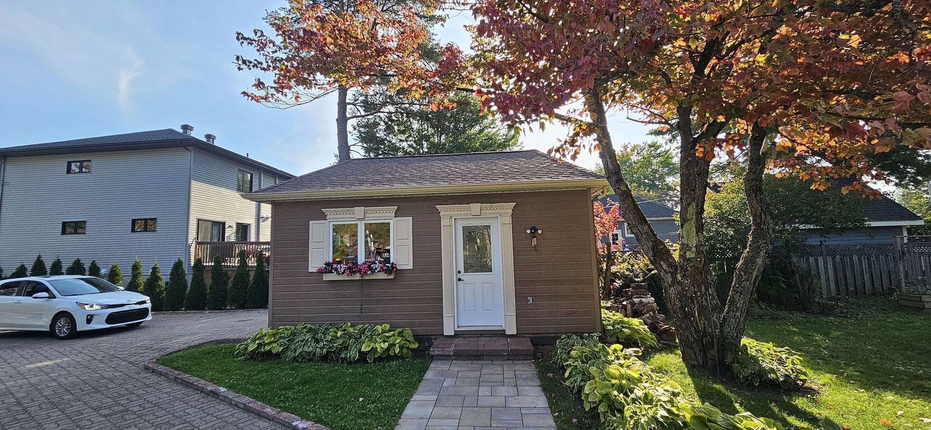 Une petite maison brune aux boiseries blanches, avec une jardinière et une allée en pierre, située à côté d'une maison moderne et d'une voiture blanche.