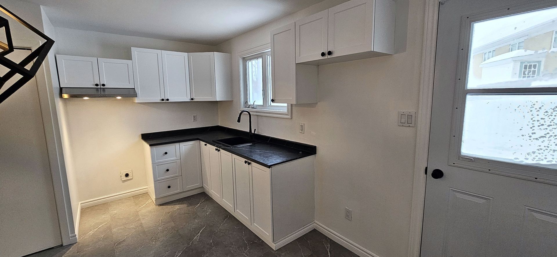 Une cuisine moderne et lumineuse comprend des armoires blanches, des comptoirs noirs, un évier et une porte donnant sur un extérieur enneigé.