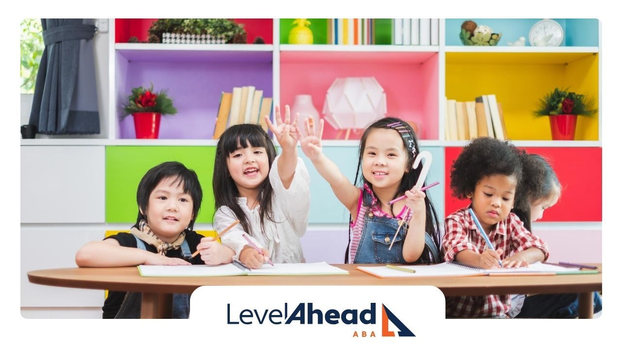 A group of children are sitting at a table with their hands in the air.