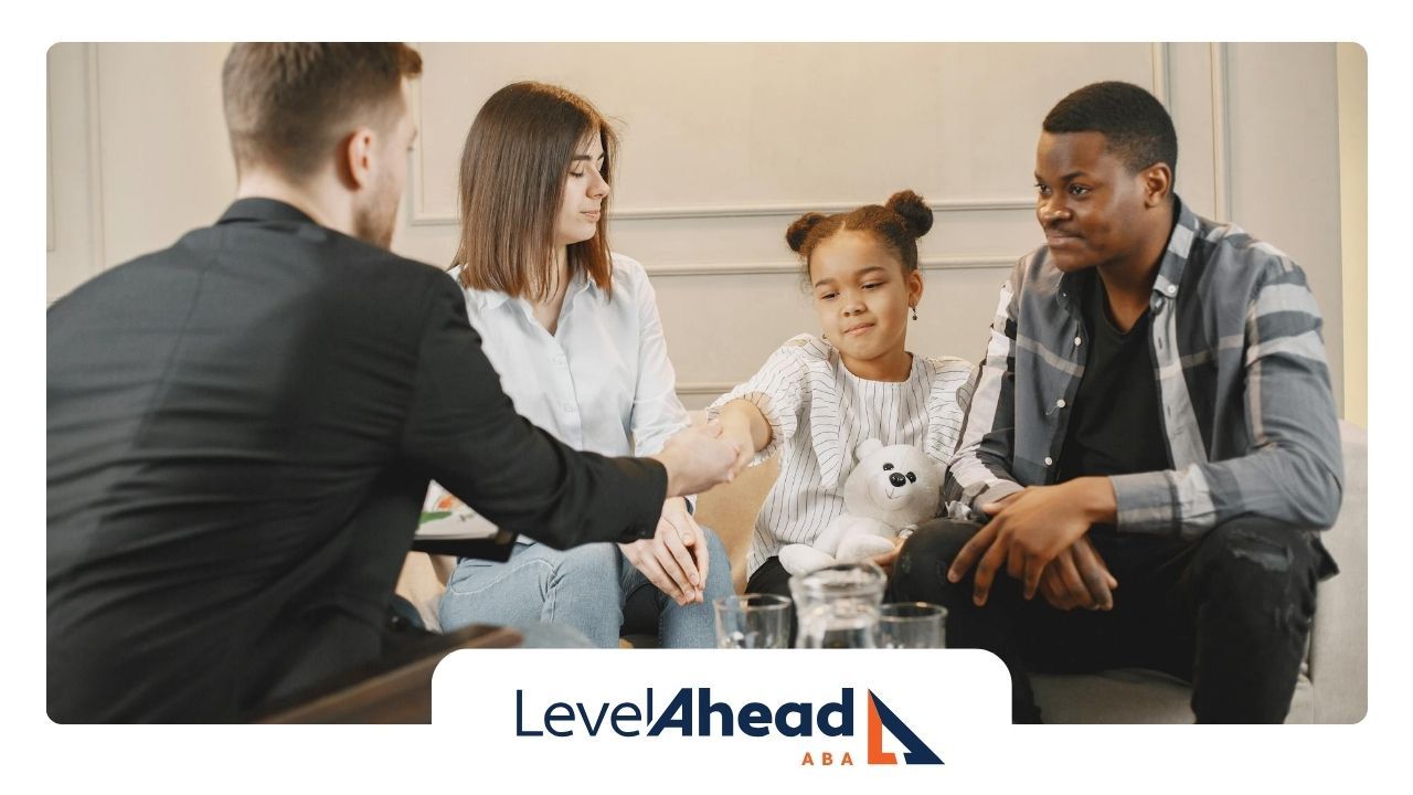 A family sitting with an ABA therapist, an autistic girl shaking hands while holding a bear in NE.