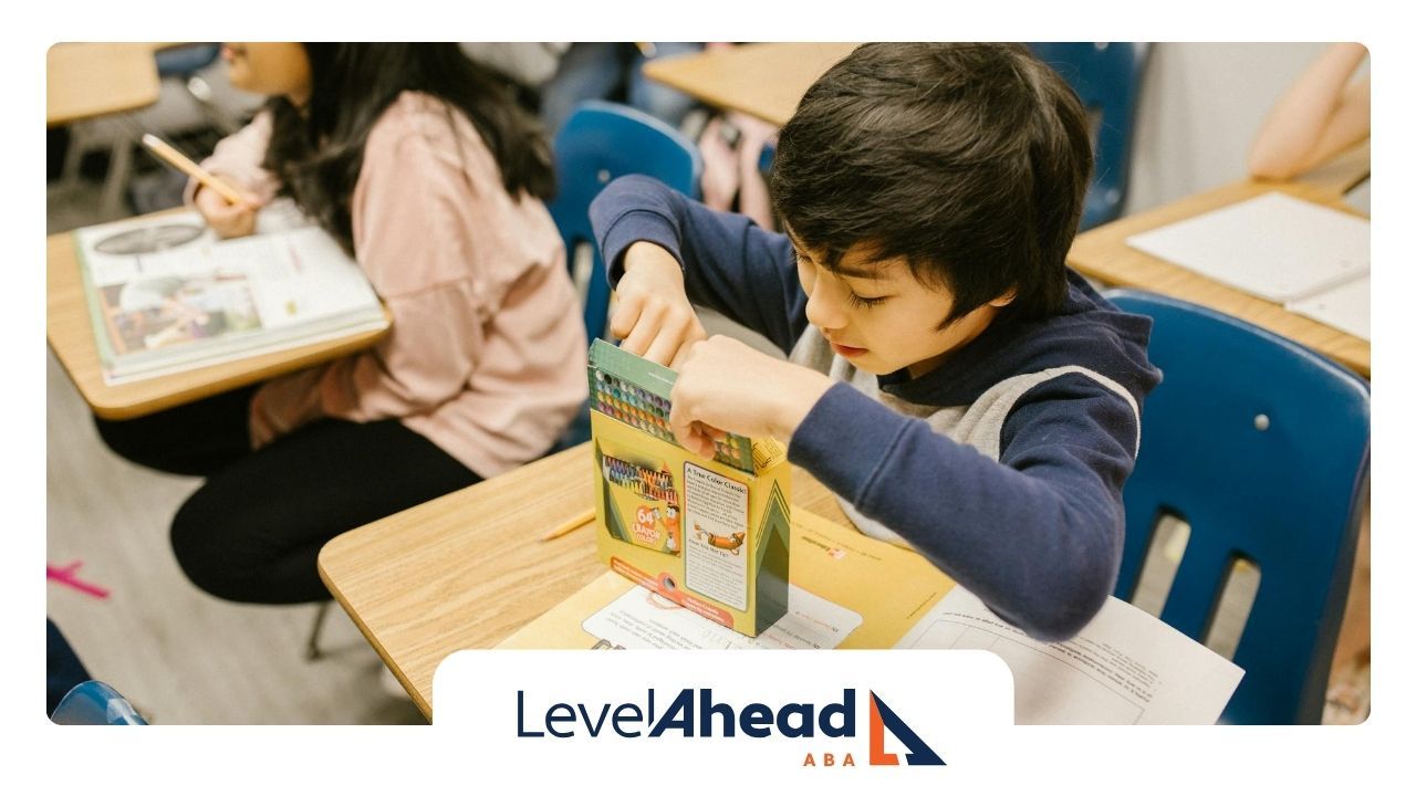 An autistic boy, focused on opening a box of crayons while surrounded by classmates in Georgia.