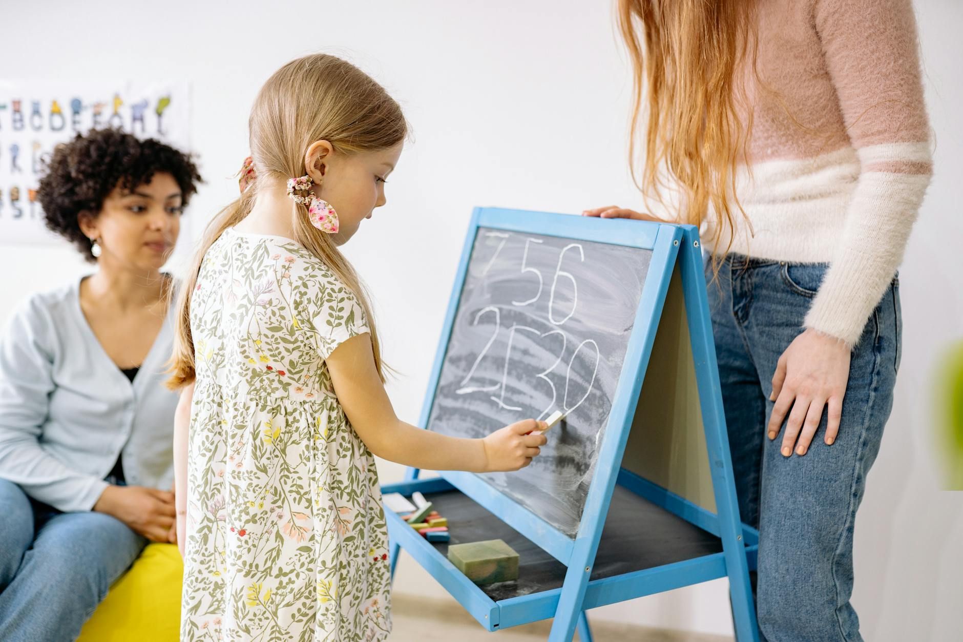 A little girl drawing on a chalkboard while a woman watches