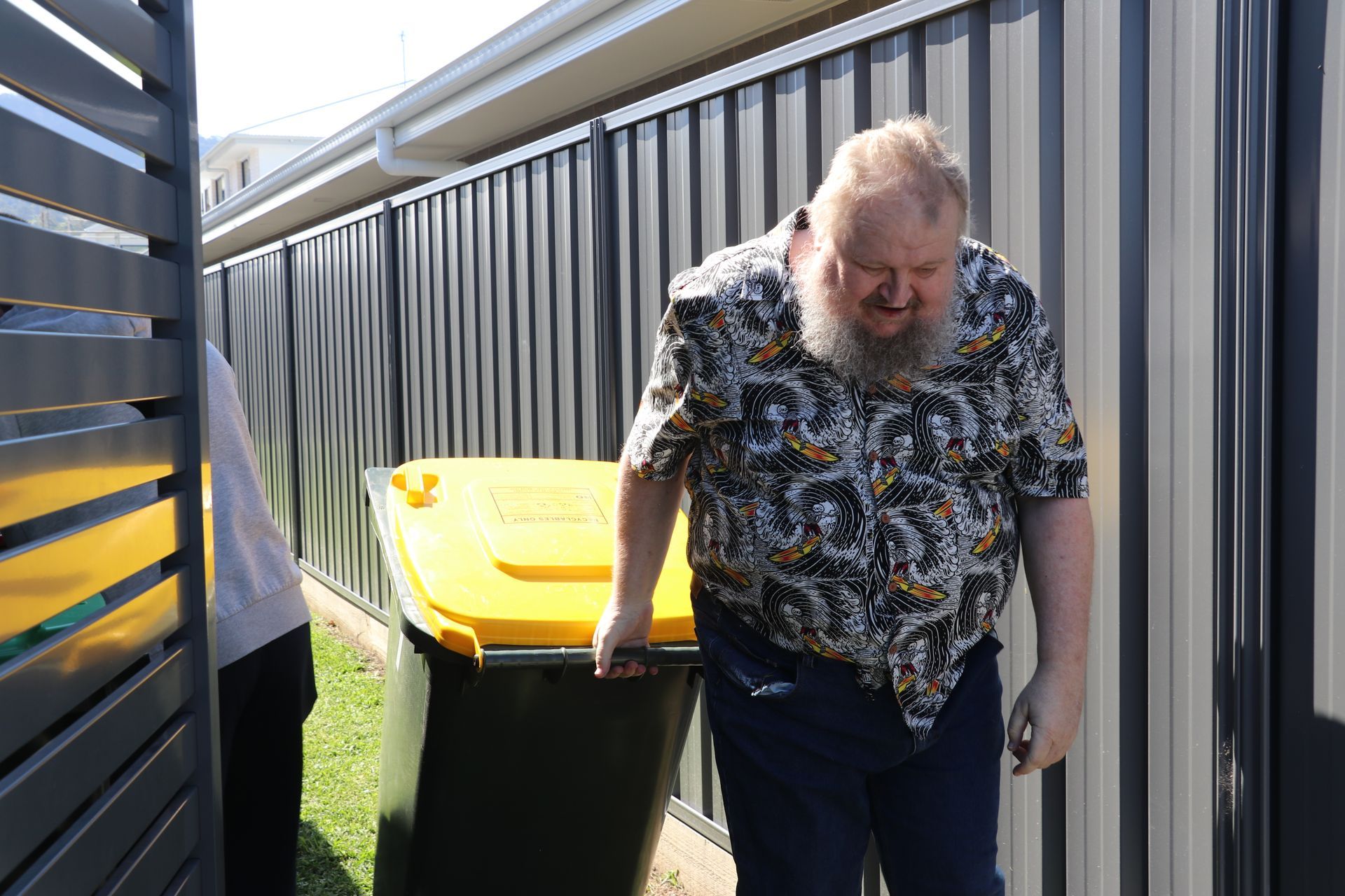 A man with a beard is pushing a yellow garbage can.