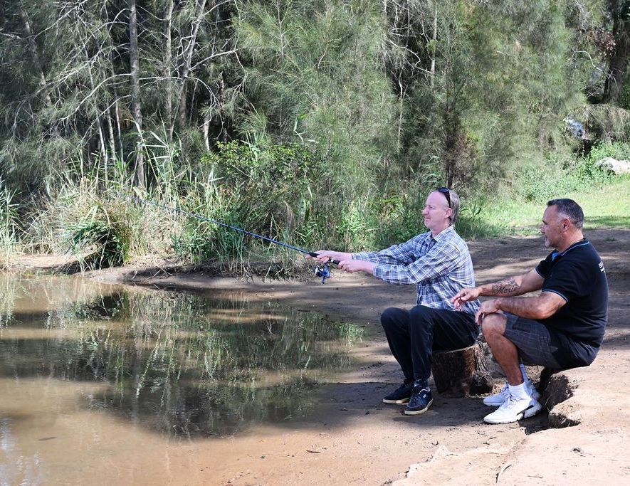 Two men are sitting on the shore of a river fishing.