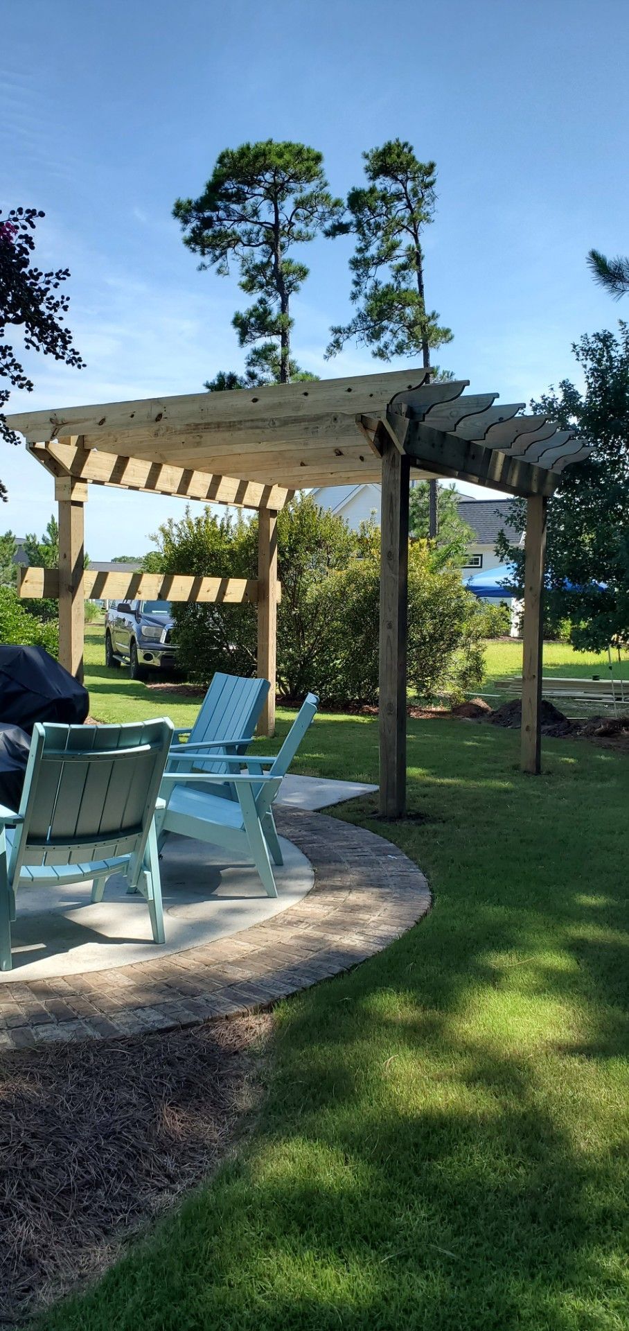 A pergola with chairs and a grill in a backyard.