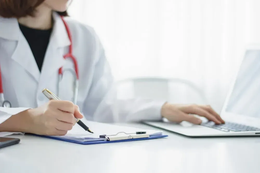 A Doctor Is Writing on A Clipboard While Using a Laptop Computer — Health Hub Mackay in Mackay, QLD