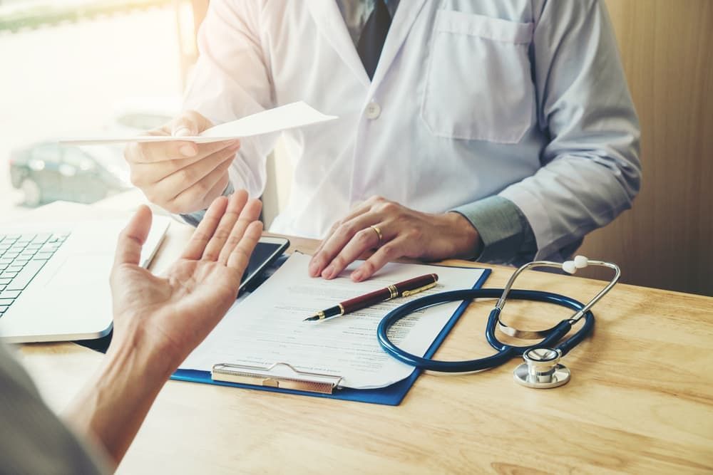 A Doctor Is Giving A Patient A Prescription While Sitting At A Desk — Health Hub Mackay In North Mackay, QLD
