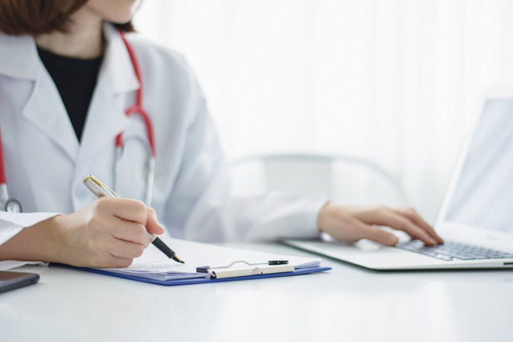 A Doctor Is Writing On A Clipboard While Using A Laptop Computer — Health Hub Mackay In Moranbah, QLD