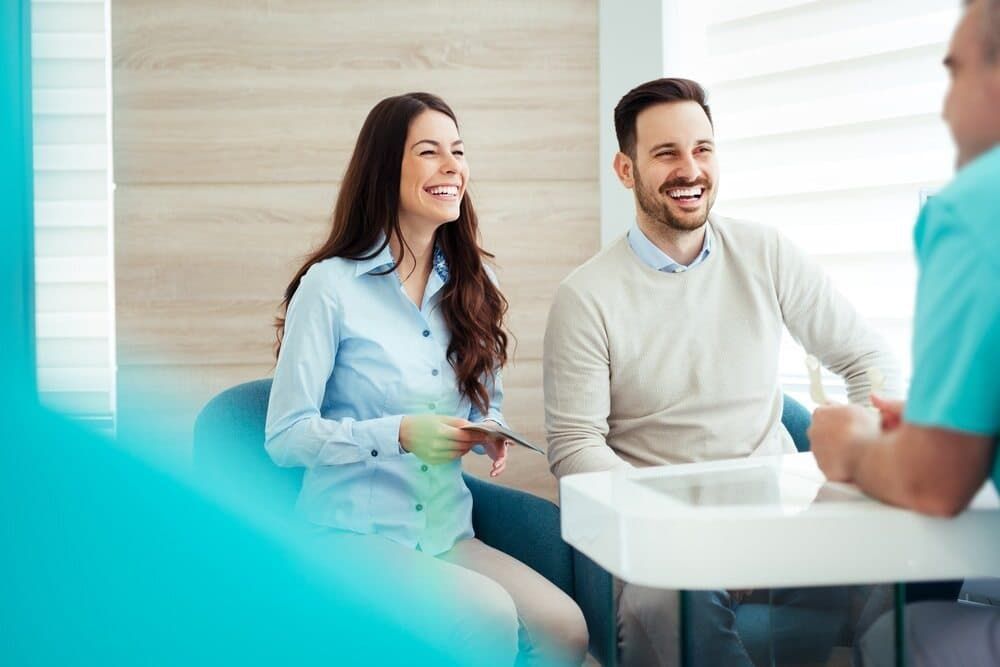 A Man And A Woman Are Sitting In A Waiting Room Talking To A Doctor — Health Hub Mackay In Proserpine, QLD