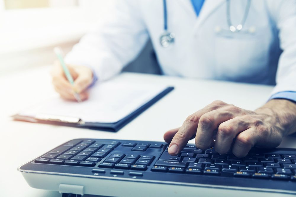 A doctor is in his office, writing on a chart with one hand and typing on a keyboard with the other.