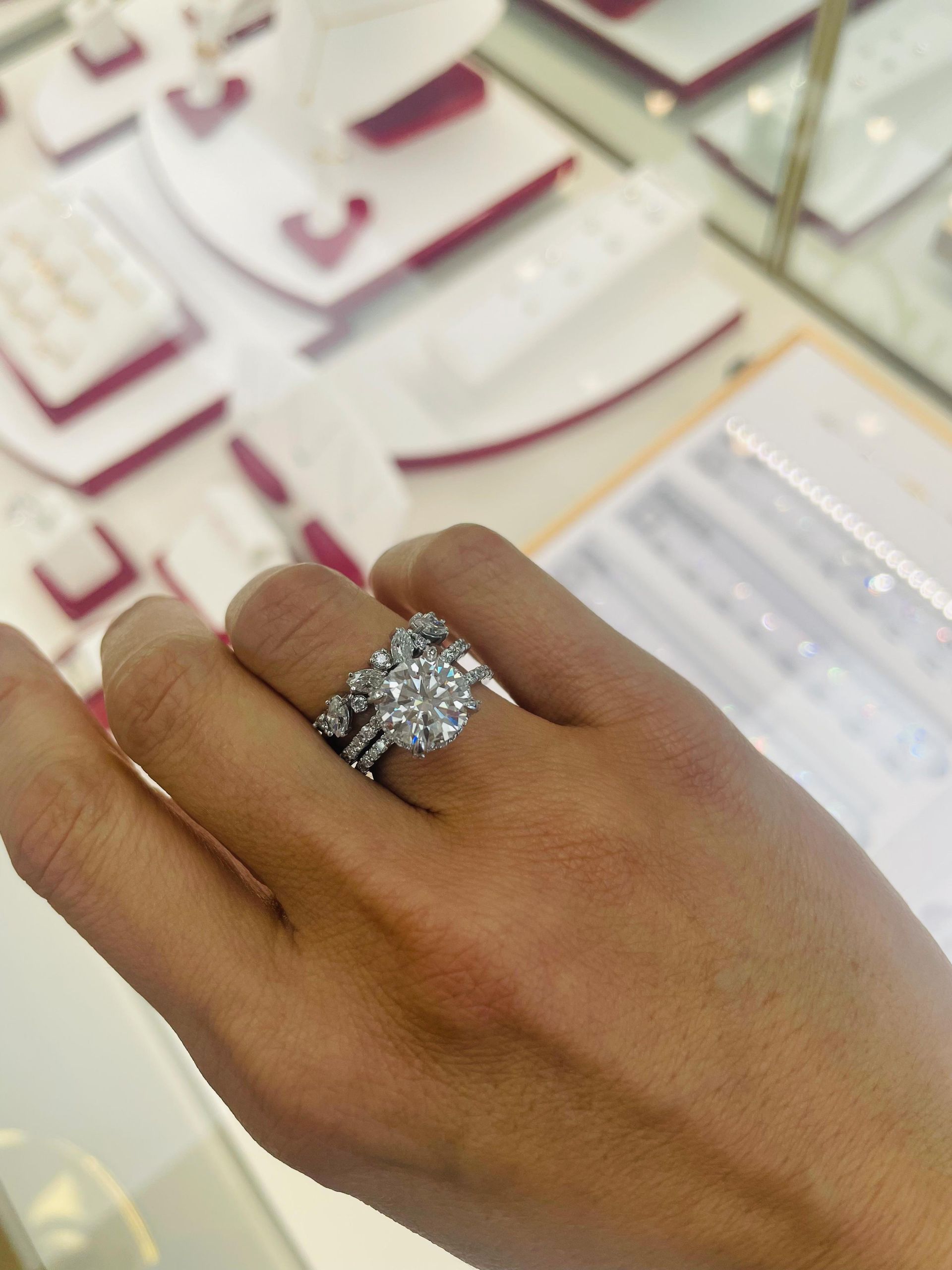 Hand wearing diamond engagement ring and band, jewelry store display in background.