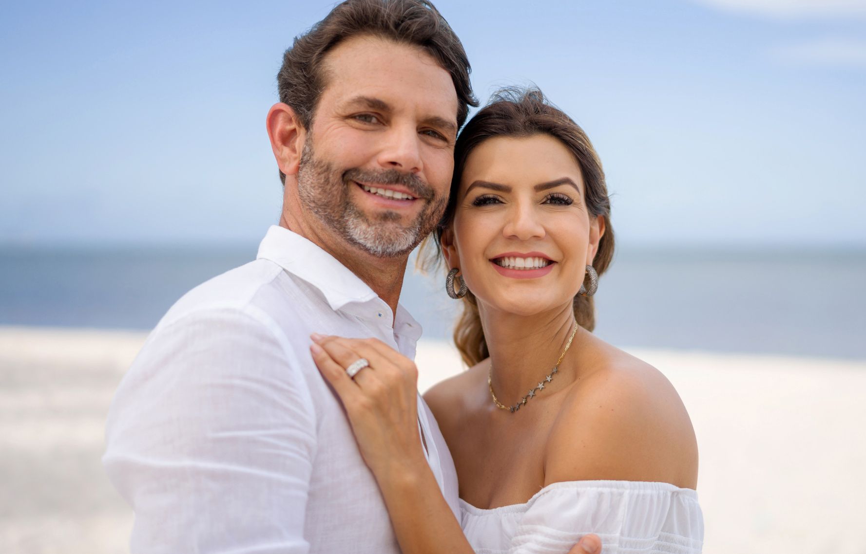 Couple smiles on a sunny beach; man has arm around woman, who displays diamond ring.