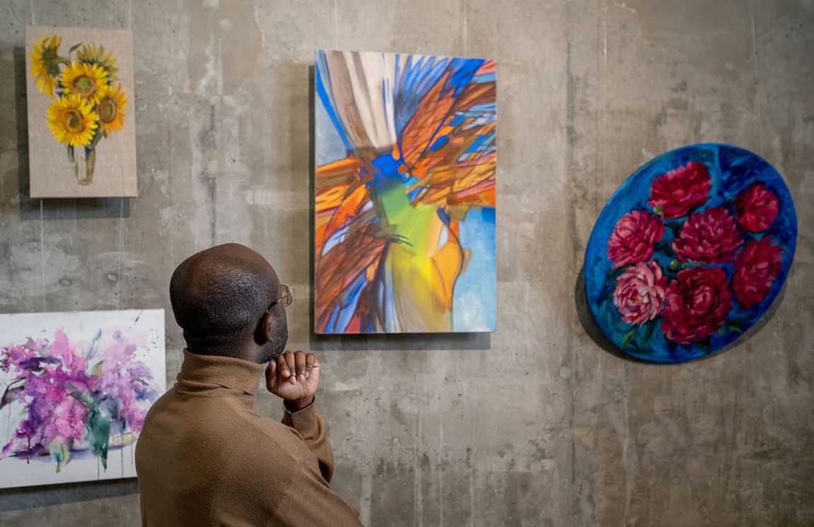 Man viewing abstract art on a gray wall. Paintings include sunflowers, floral, and colorful designs.