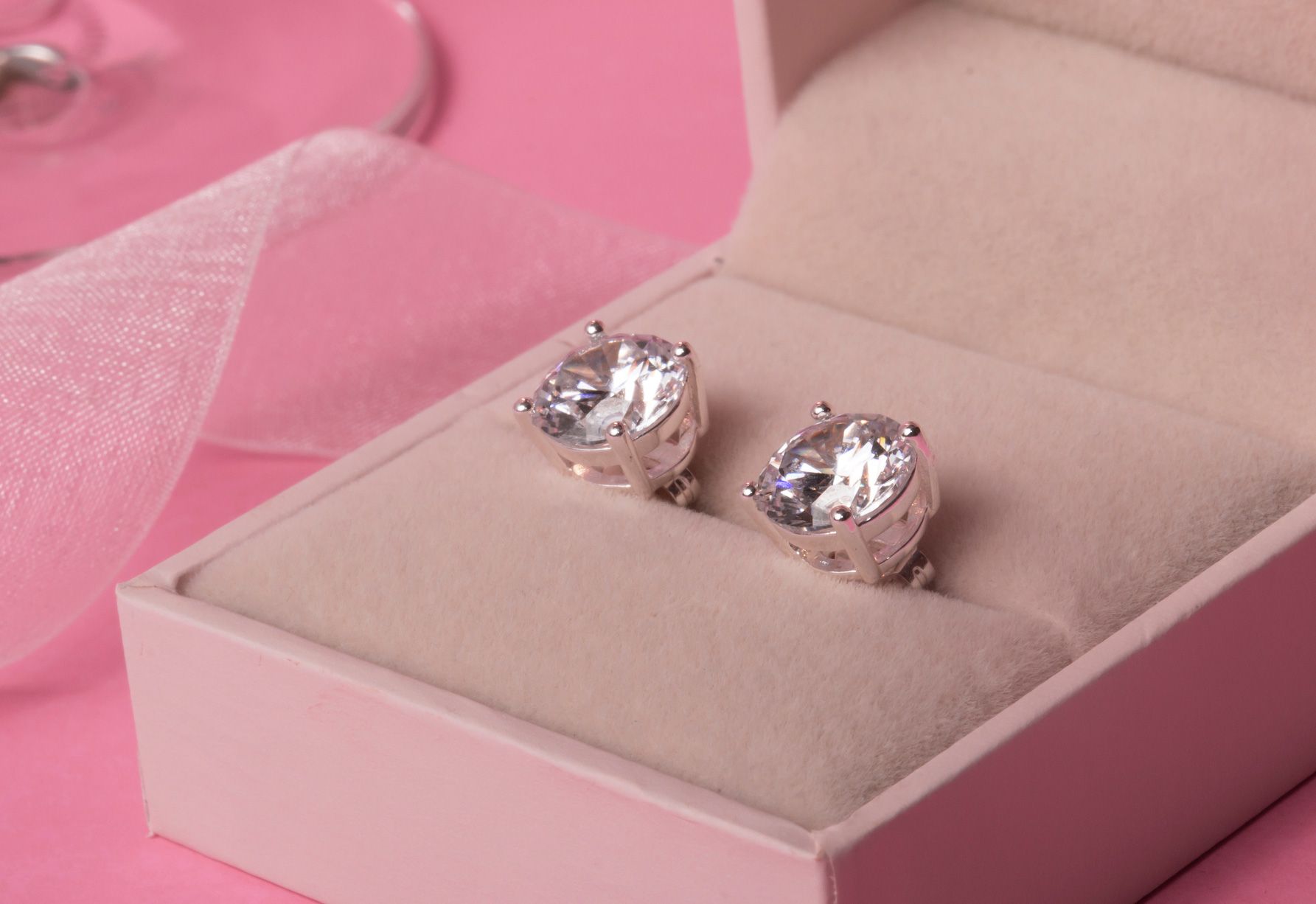 Pair of diamond stud earrings in a pink jewelry box, with pink background and ribbon.