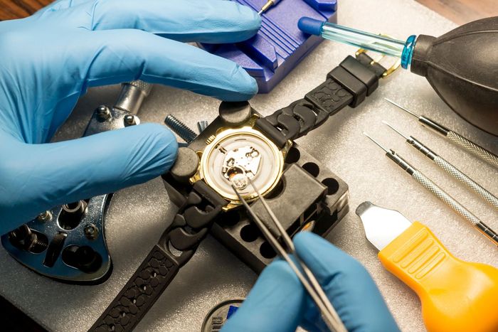 Hands in blue gloves repairing a watch using tweezers, tools, and a black holder on a white surface.