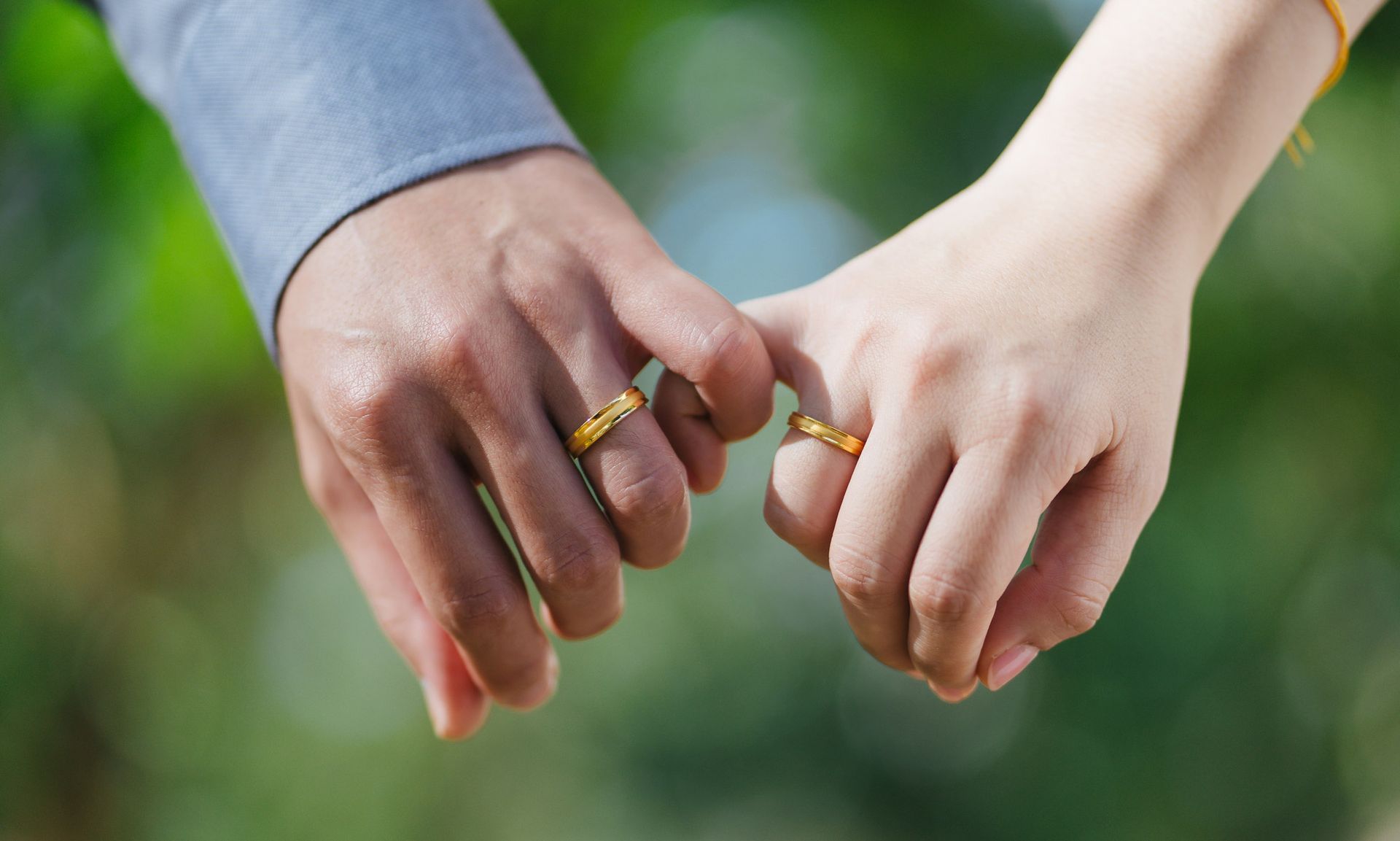 Two hands, linked pinky fingers, gold wedding bands; blurred green background.