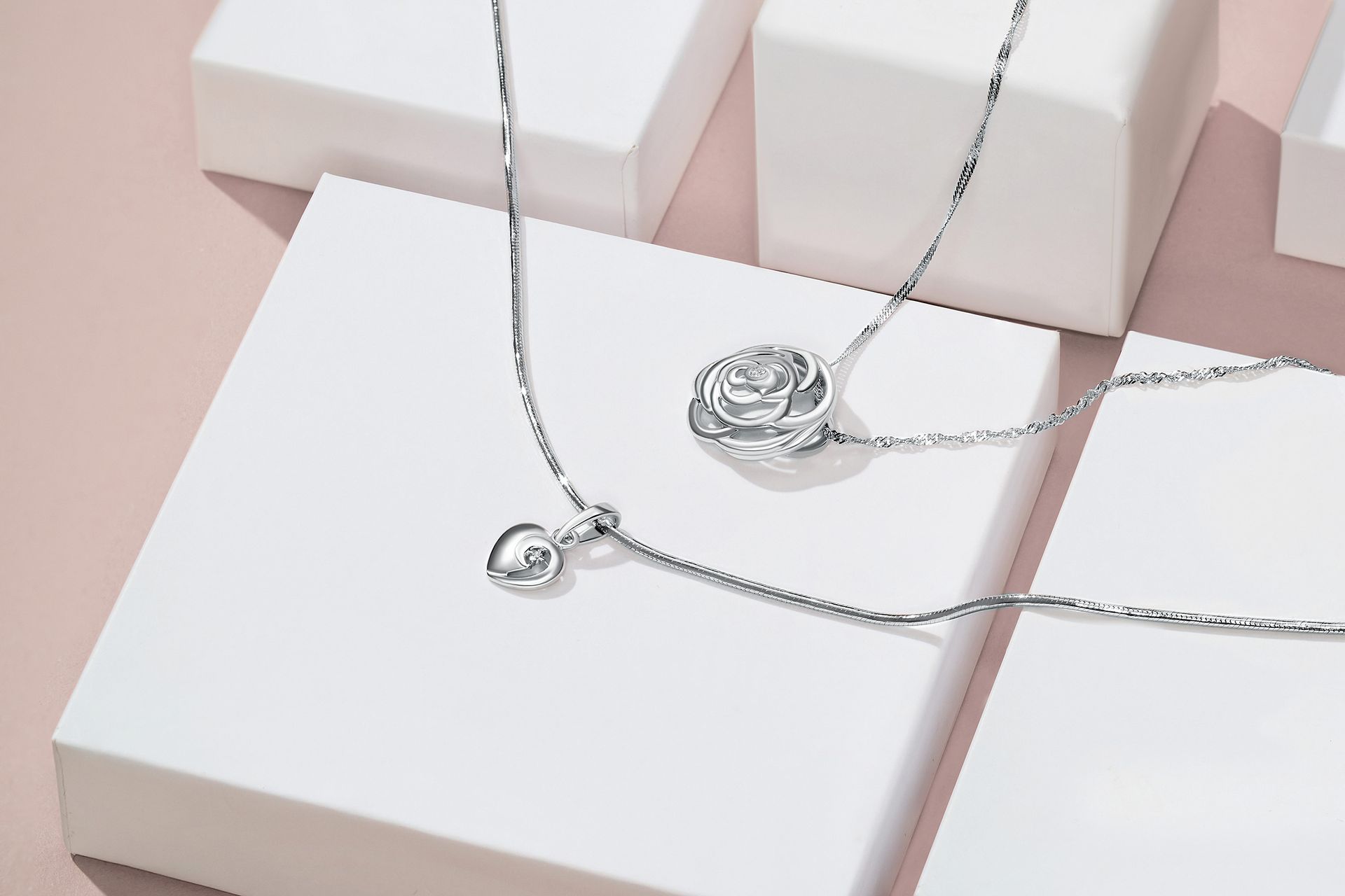 Two silver necklaces on white blocks; one with a rose pendant, one with a small charm.