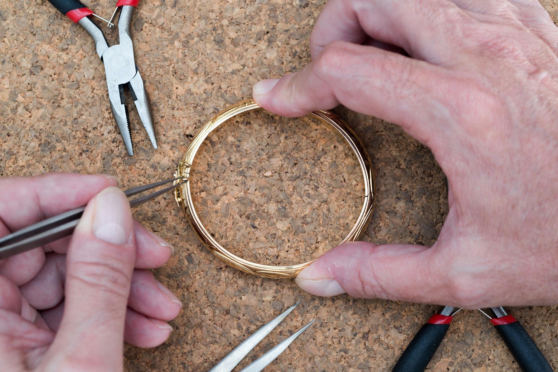 Hands repairing a gold bracelet with pliers and tweezers on a textured surface.