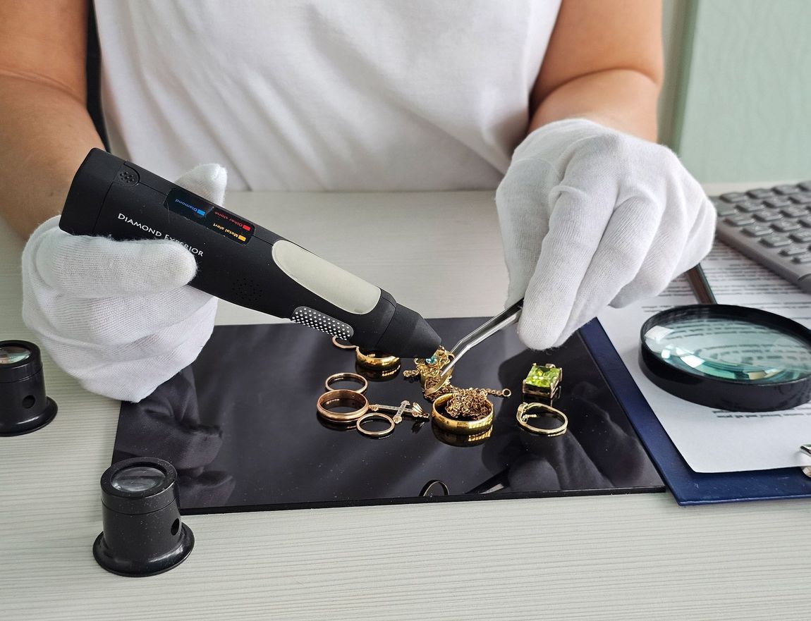 A person wearing white gloves examines jewelry with a testing device and tweezers at a desk.