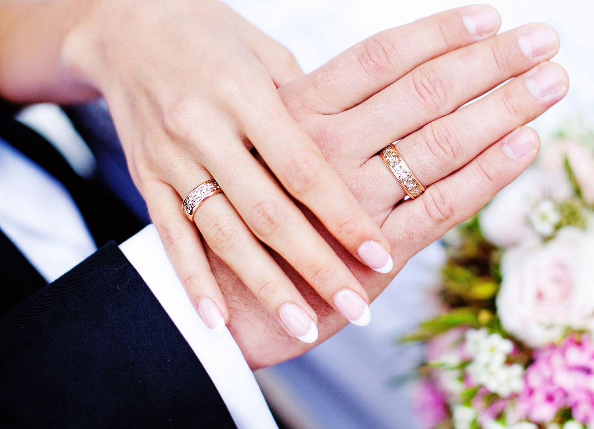 Hands of a couple with wedding rings, one hand on top of the other, near a bouquet.