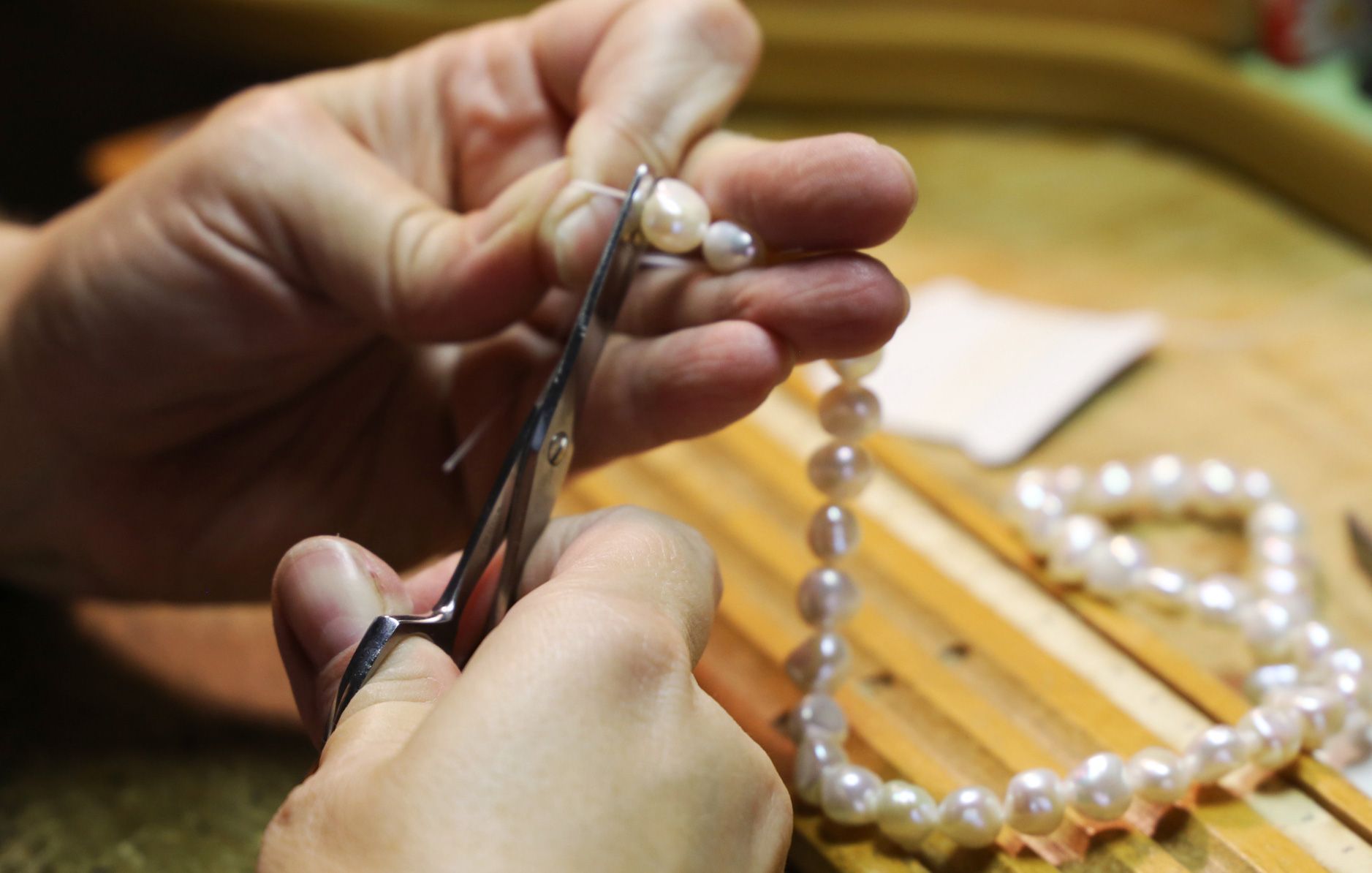 Hands cutting pearl necklace with scissors, wooden workbench.