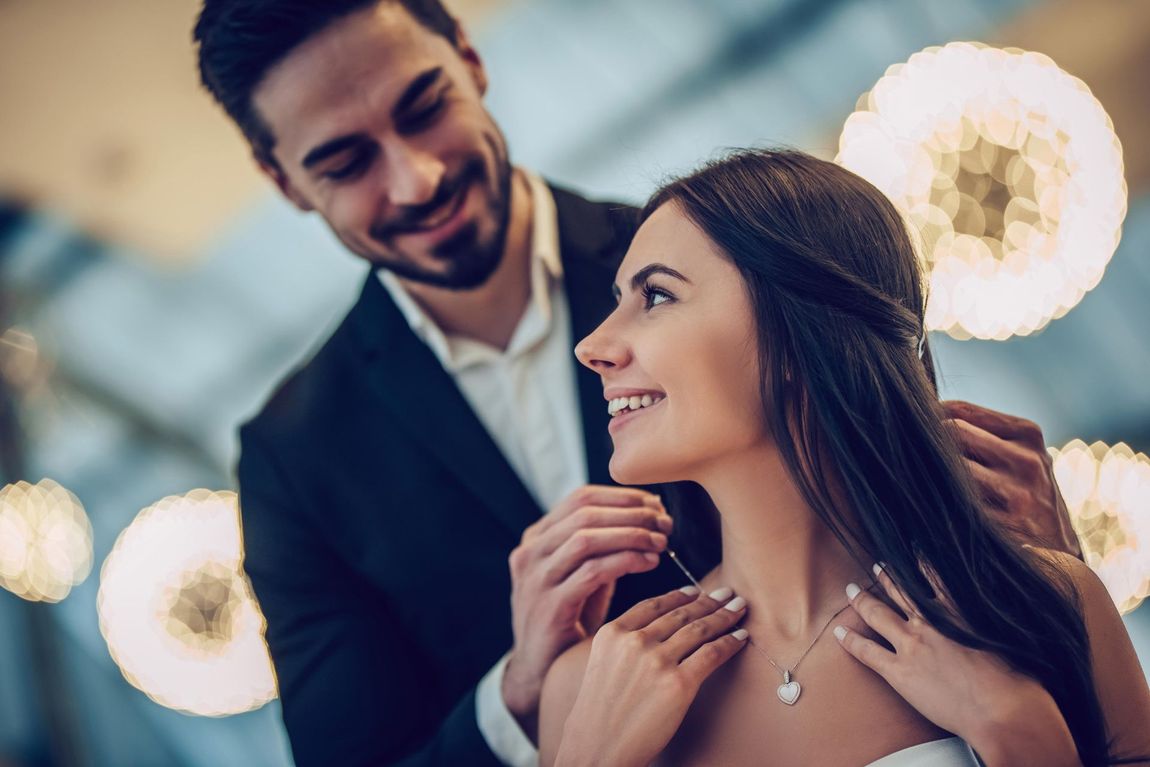 Man putting necklace on woman. They're smiling, indoors.