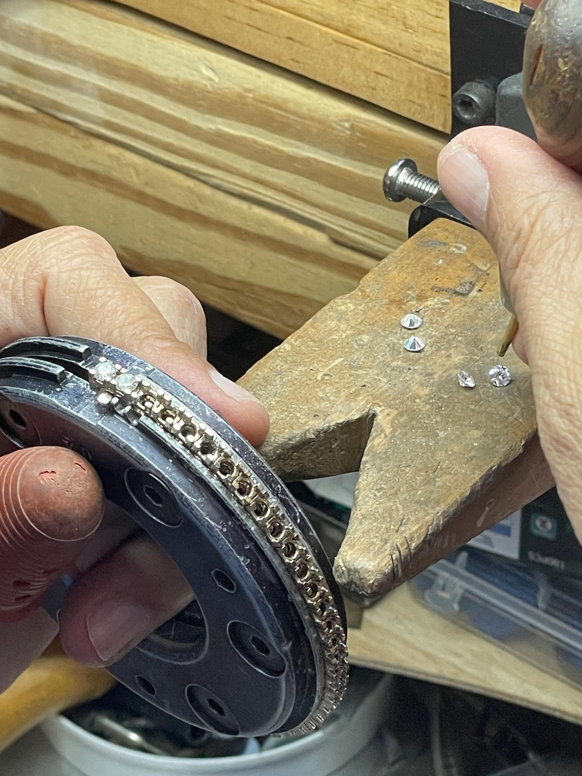 Jeweler's hands setting diamonds into a metal bracelet, using tools in a workshop.