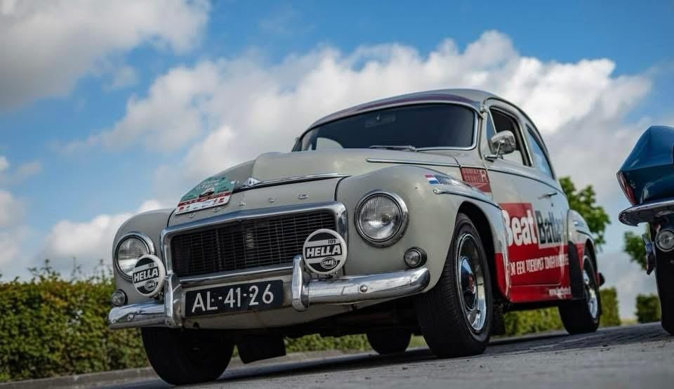 Grijze oldtimer Volvo met rode race-stickers en kentekenplaat AL-41-26, buiten onder een bewolkte hemel.