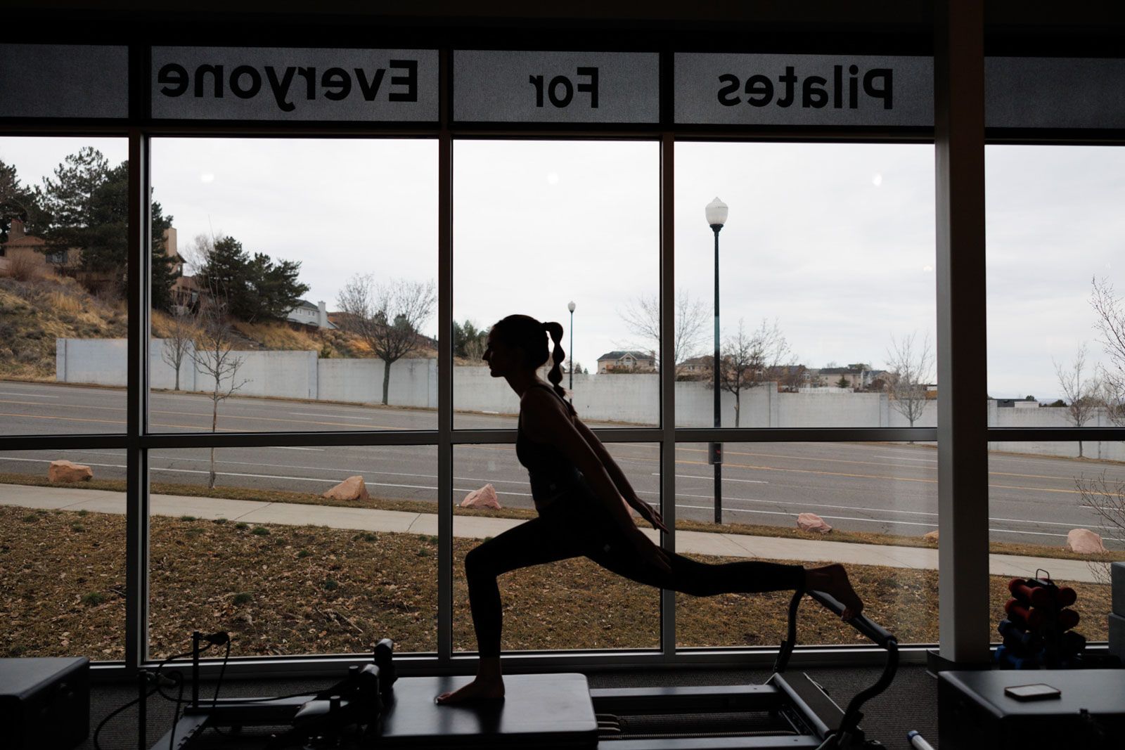 Silhouette of a person stretching in a gym by large windows, with “Pilates for Everyone” signage above.