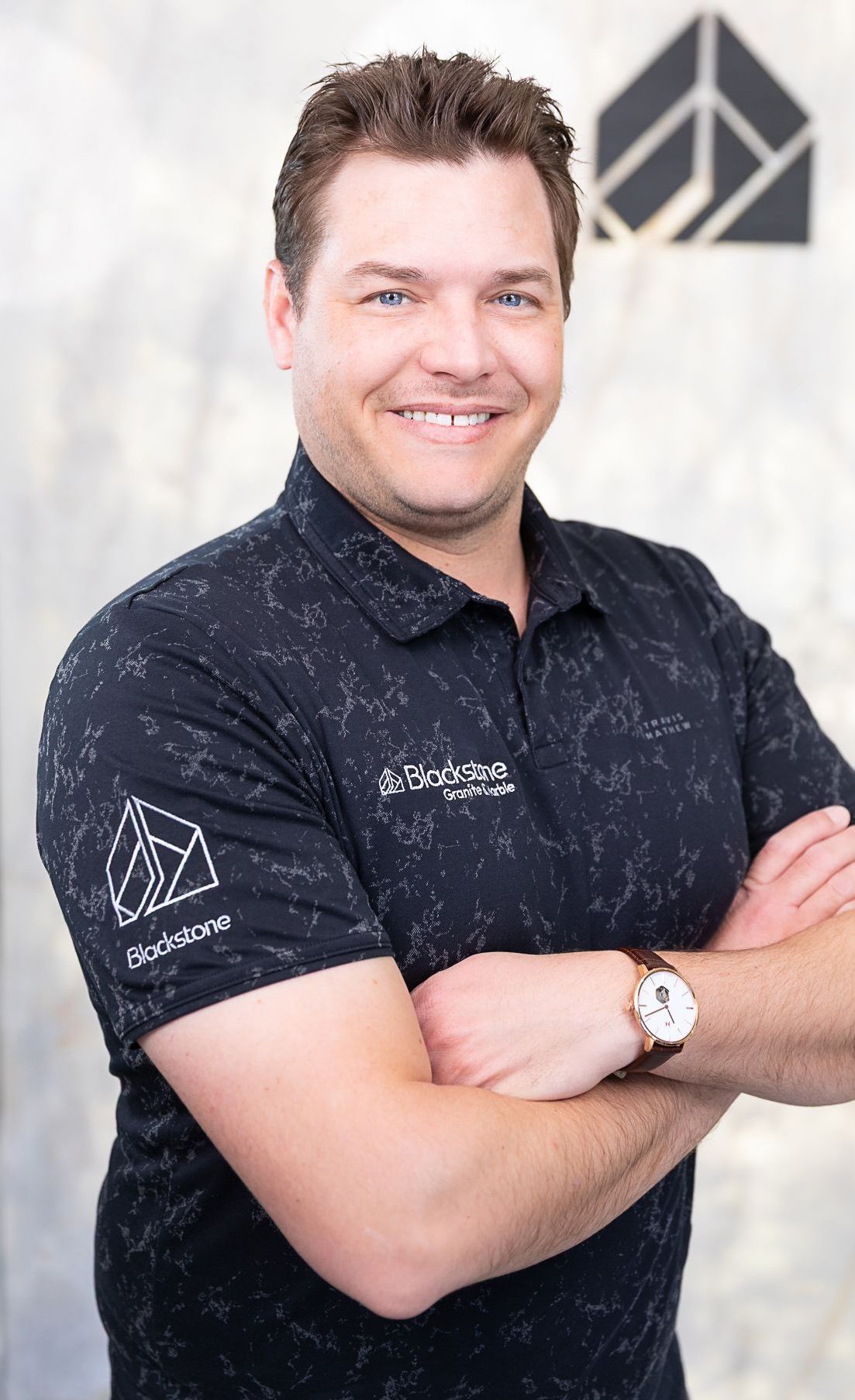 A man wearing a black shirt and a watch is standing with his arms crossed.