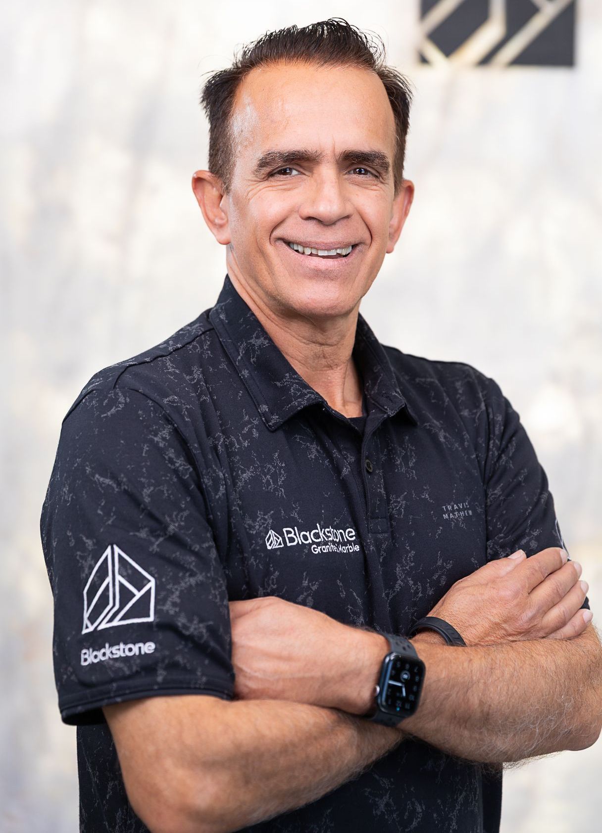 A man in a black shirt is standing with his arms crossed and smiling.