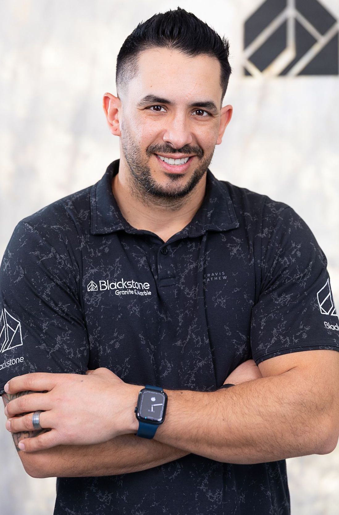A man wearing a black polo shirt and a watch is standing with his arms crossed.