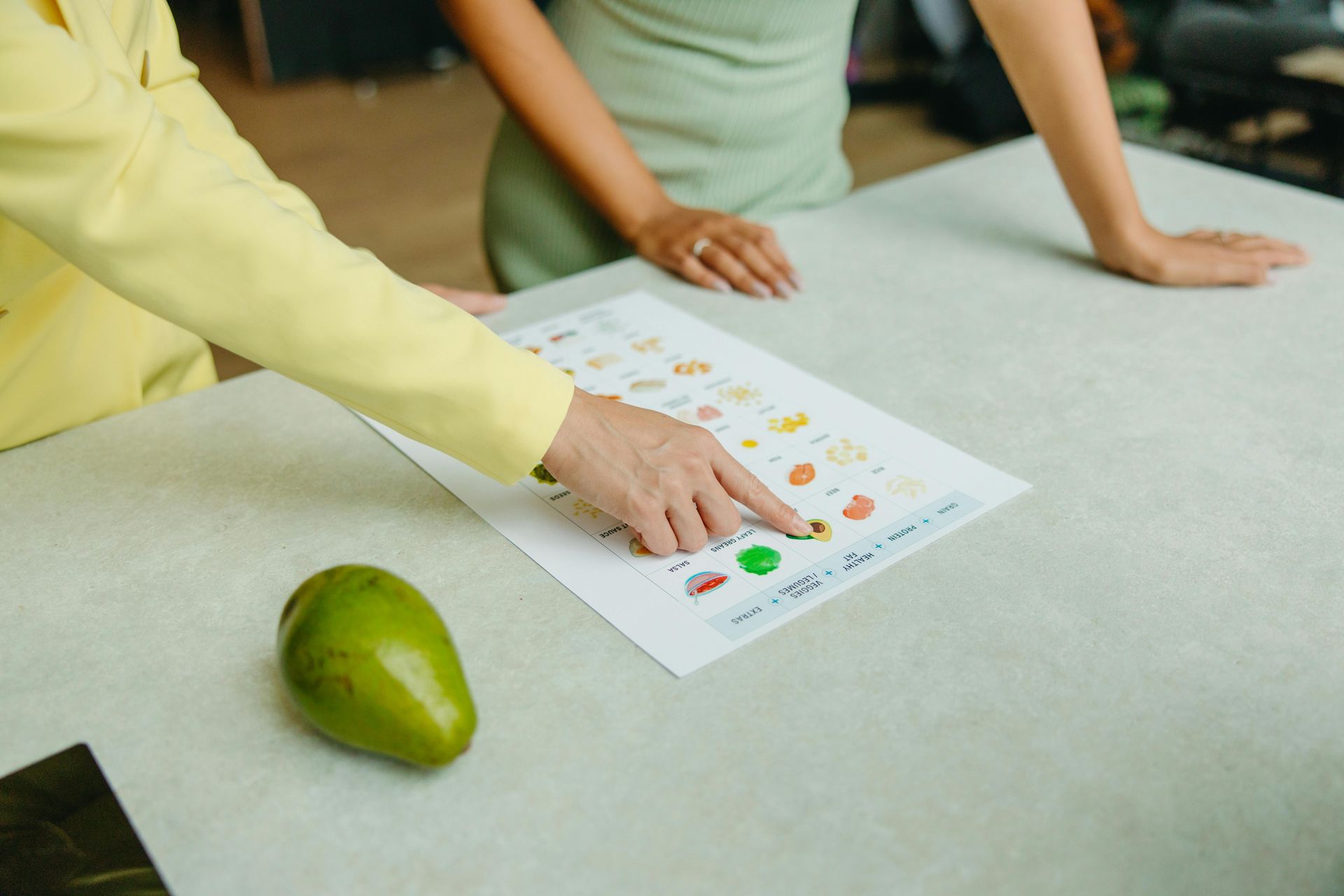Two people pointing at a chart with food icons, next to an avocado on a table.