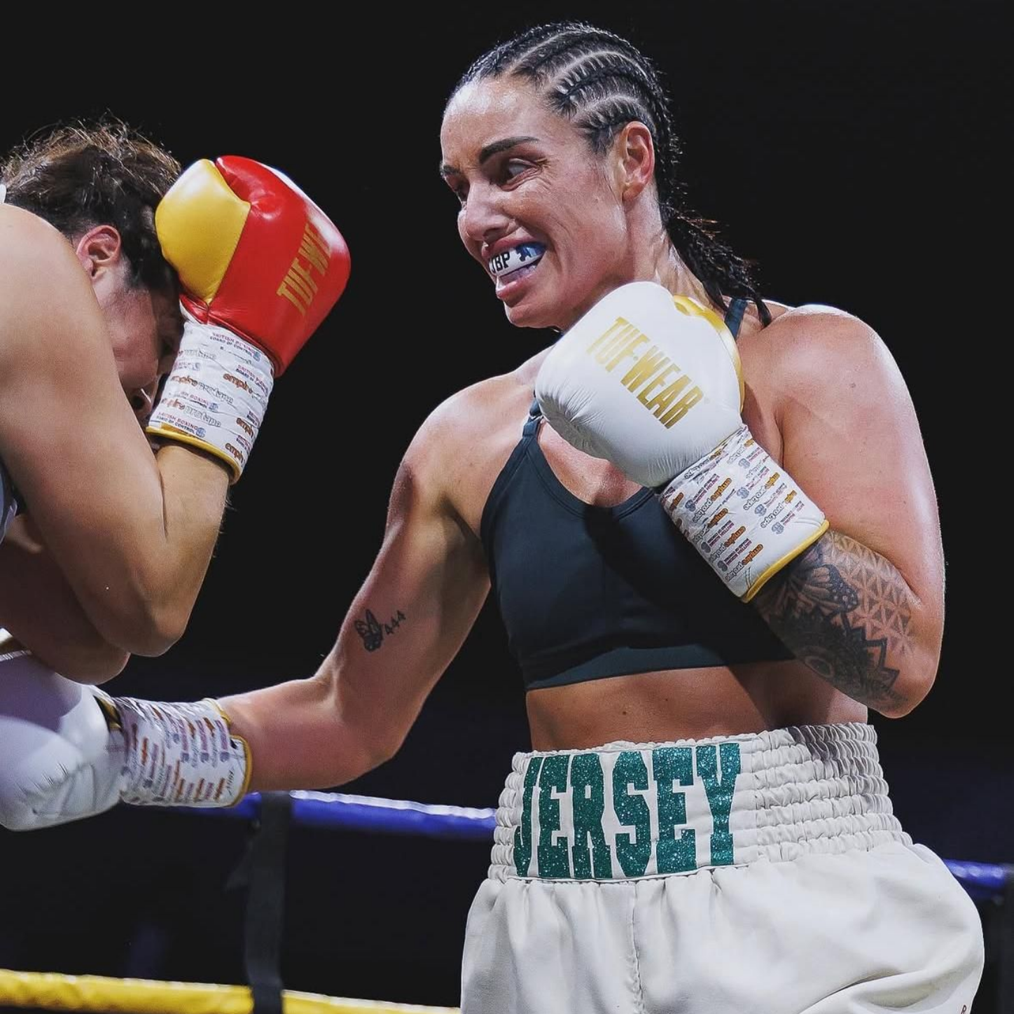 Boxer in the ring, throwing a punch. She wears white gloves, a sports bra, and boxing shorts labeled 