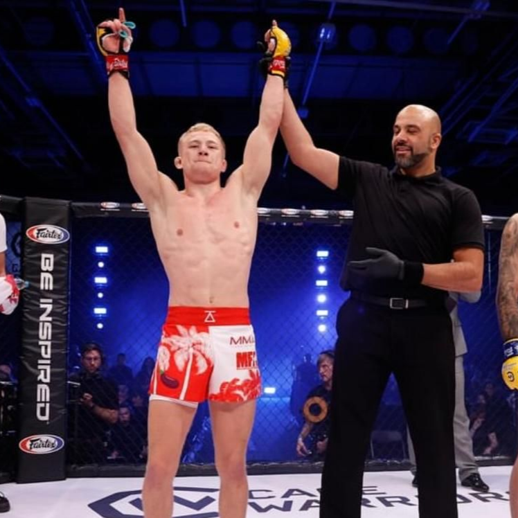 MMA fighter in red shorts raises arms in victory inside the ring with a referee.