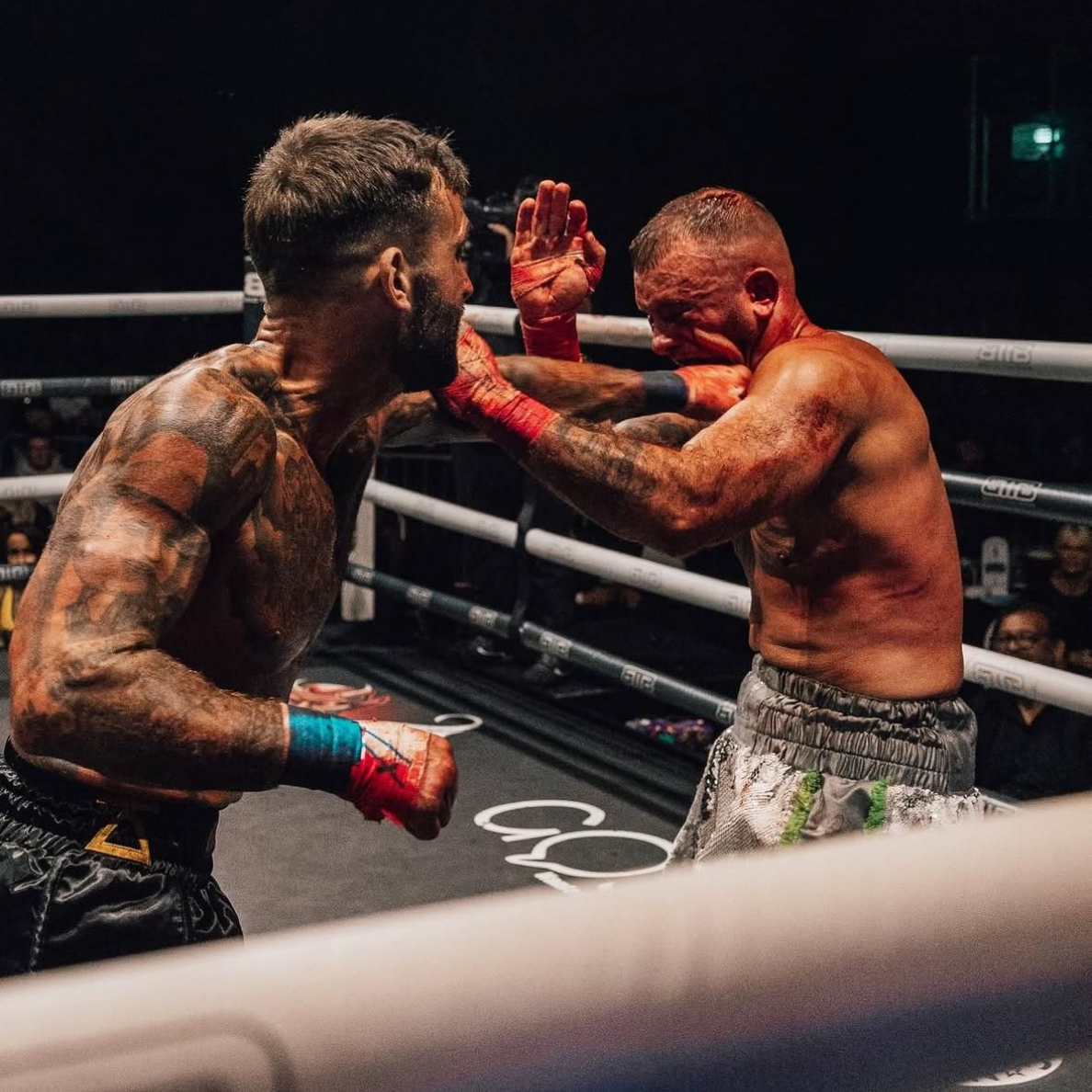 Boxers in a ring, one punches the other. Bloodied face, tattoos, ring ropes, and crowd.