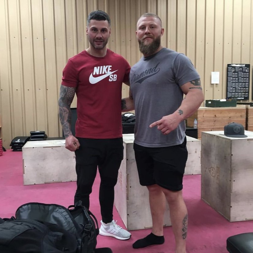 Two men pose in gym; one in red Nike shirt, the other in gray, with weight boxes and exercise gear.
