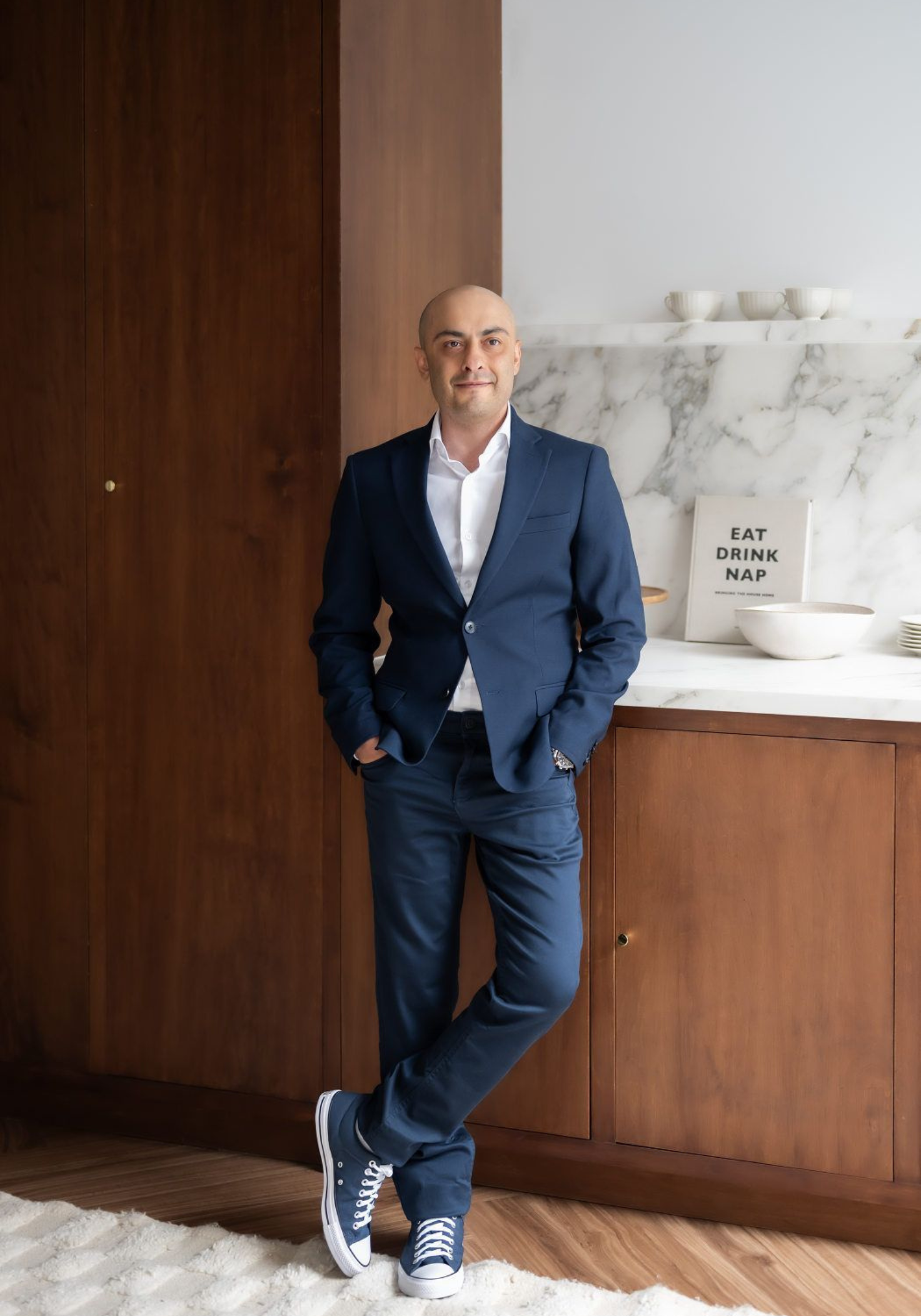 A man in a suit and sneakers is leaning against a wall in a kitchen.