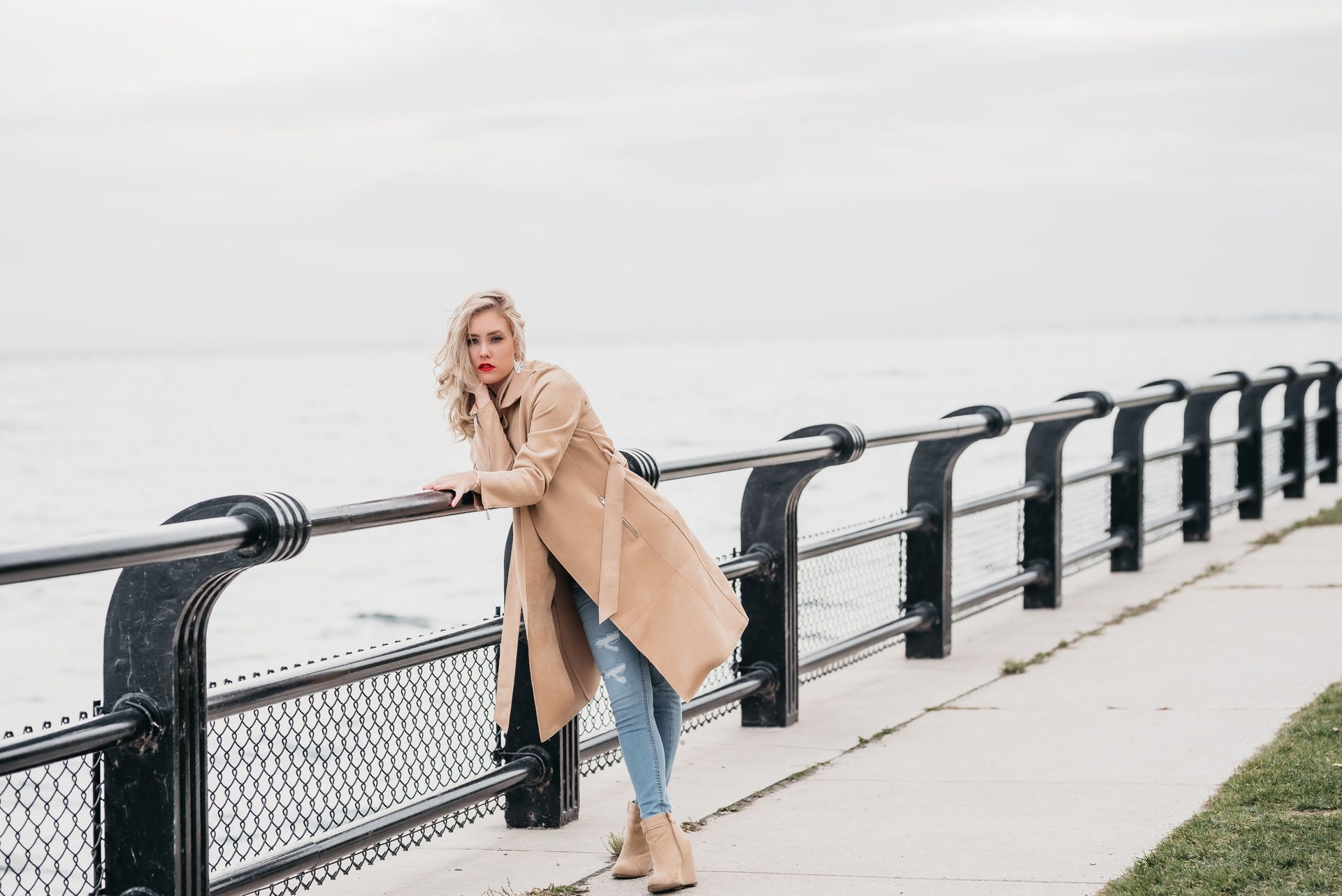 A woman in a trench coat is leaning on a railing near the water.