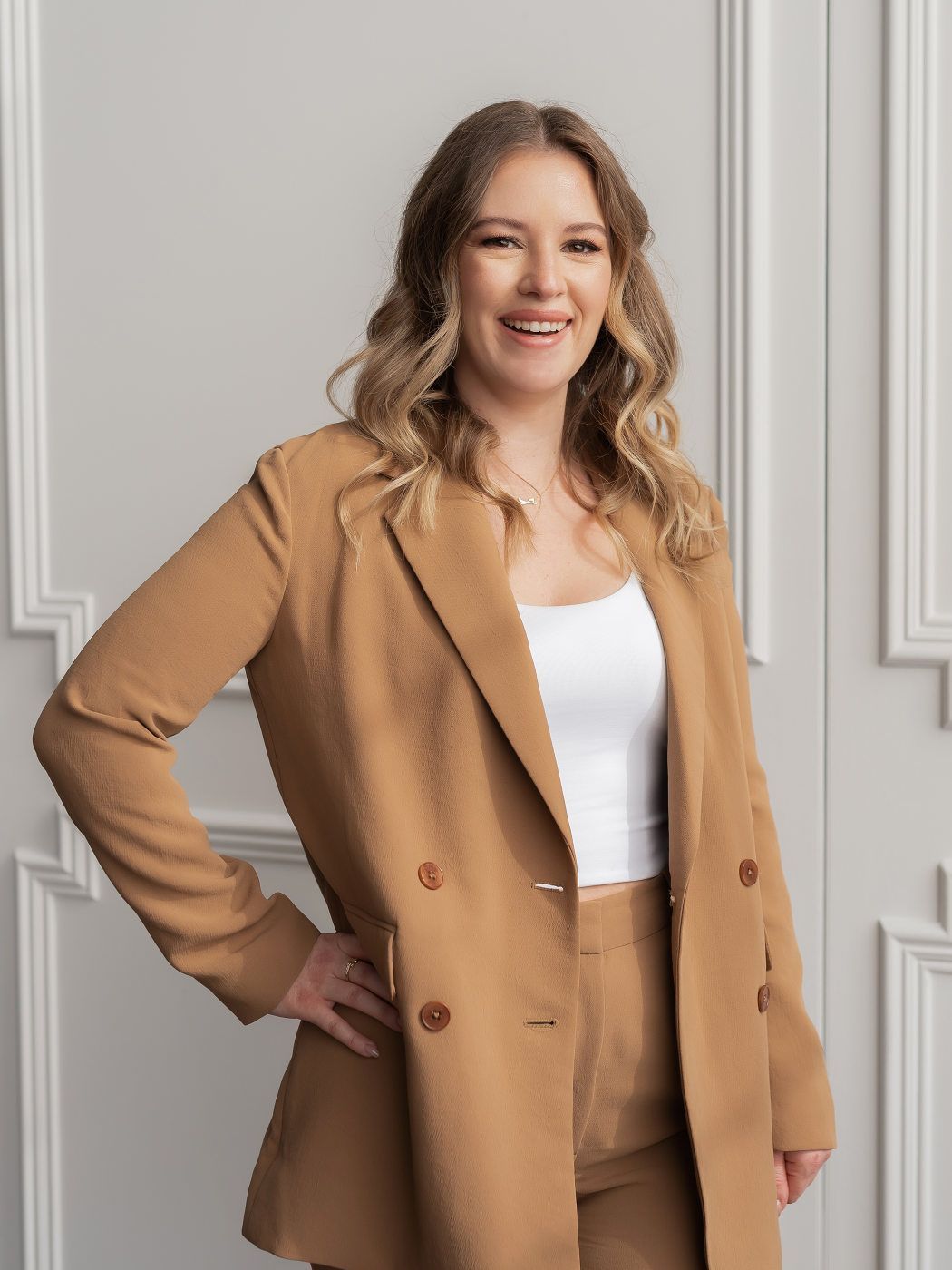 A woman in a brown suit is standing in front of a white wall.