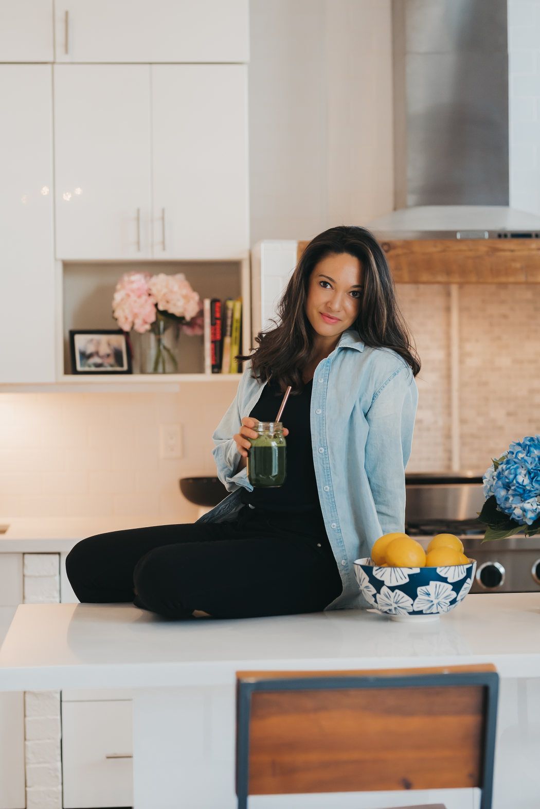 A woman is sitting on a kitchen counter drinking a green smoothie.