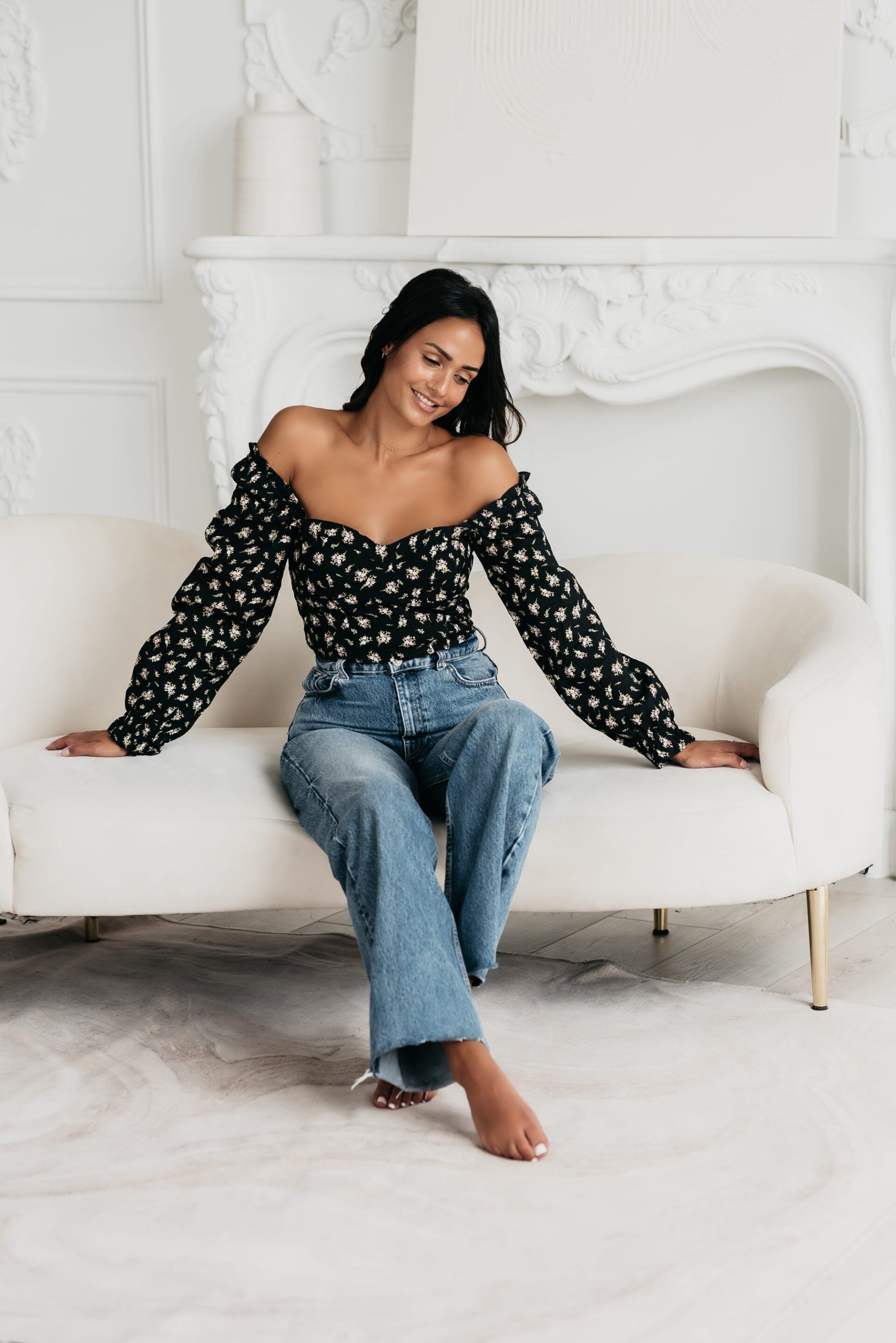 A young professional wearing a black off shoulder top and jeans sitting in an all-white luxury lounge set up