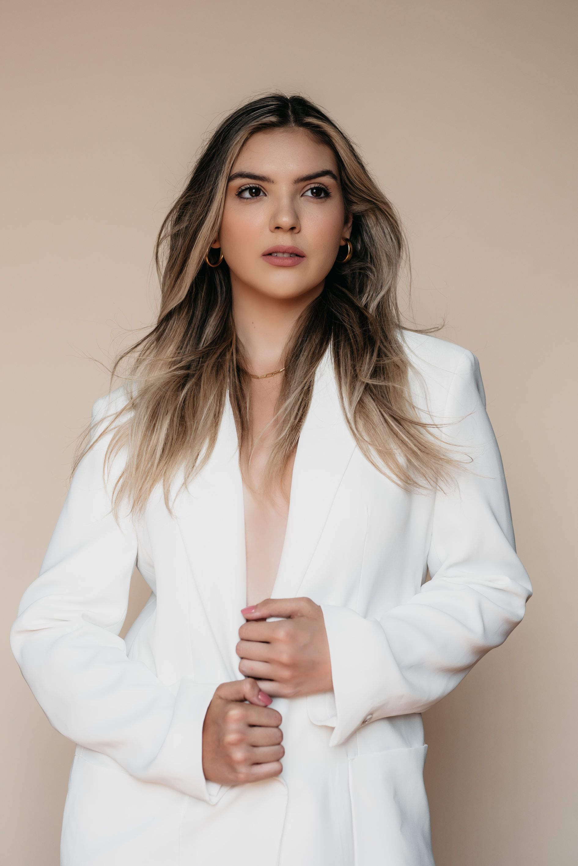 A stylish woman with long hair wearing a white blazer, standing against a beige background.