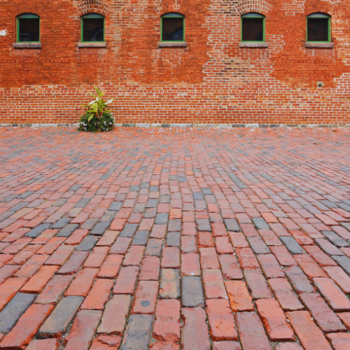 Cobblestone pathway in front of a red brick wall with green-framed windows and a small plant.