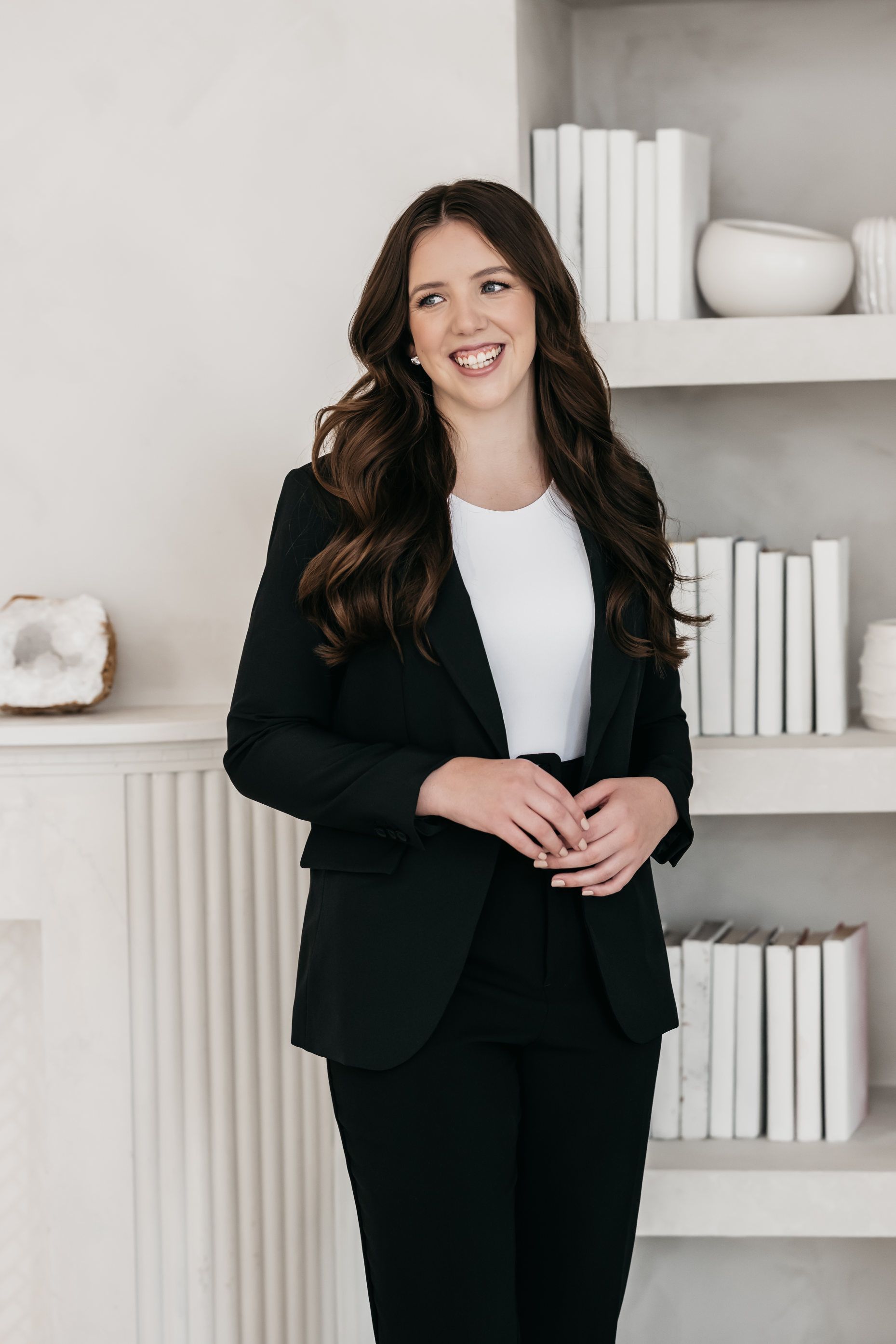 A woman in a black suit is standing in front of a bookshelf.
