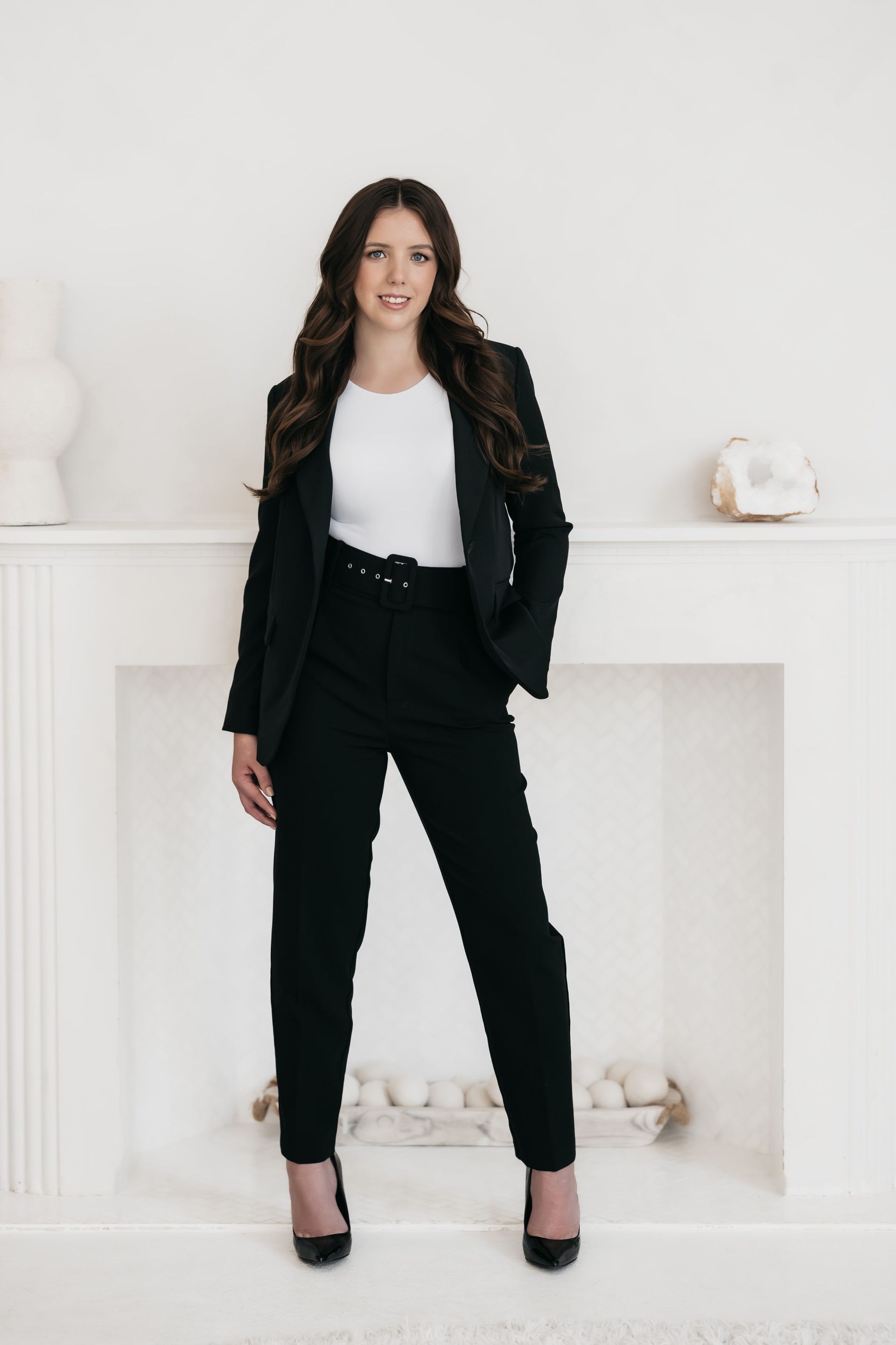 A woman posing for corporate headshots in front of a fireplace, dressed in a sophisticated black blazer and heels.