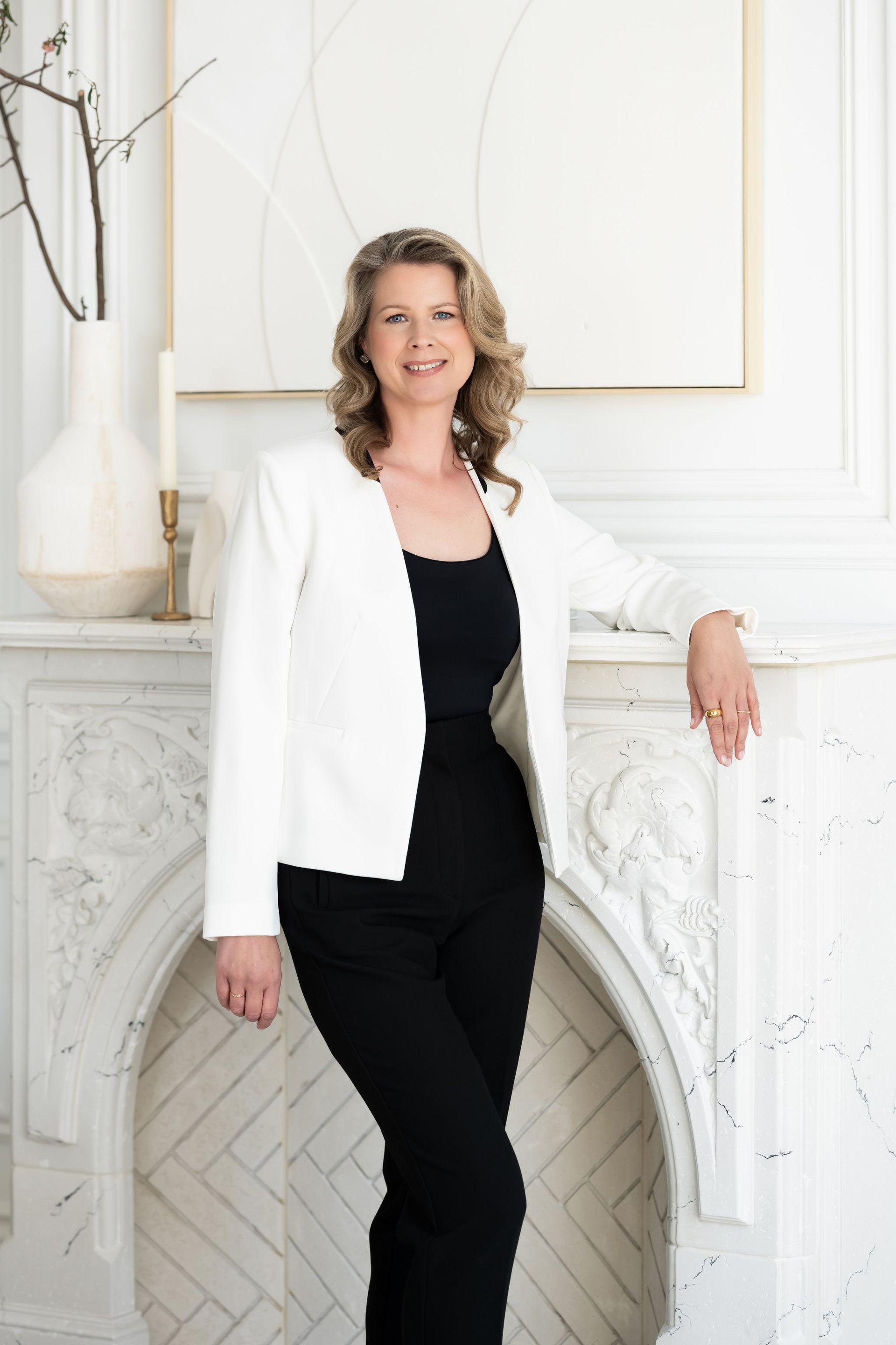 A woman in a white jacket and black pants is standing in front of a fireplace.