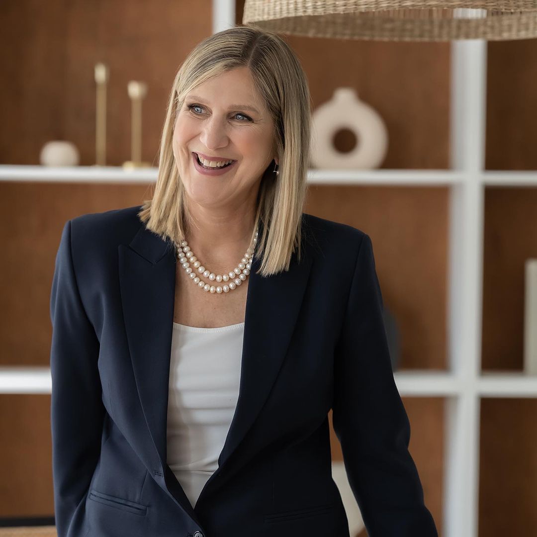 Realtor in a navy blazer with a pearl necklace, smiling in front of office shelves.