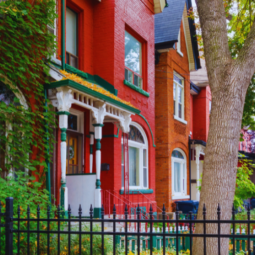 Red brick row houses with green and white accents, surrounded by lush greenery and a black iron fence.